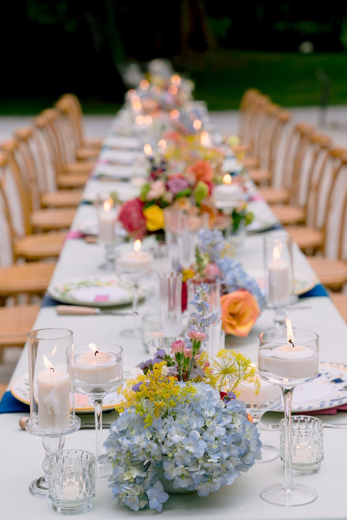 A long outdoor dining table is set with lit candles, glassware and bright floral centrepieces including blue hydrangeas, with wooden chairs along both sides.
