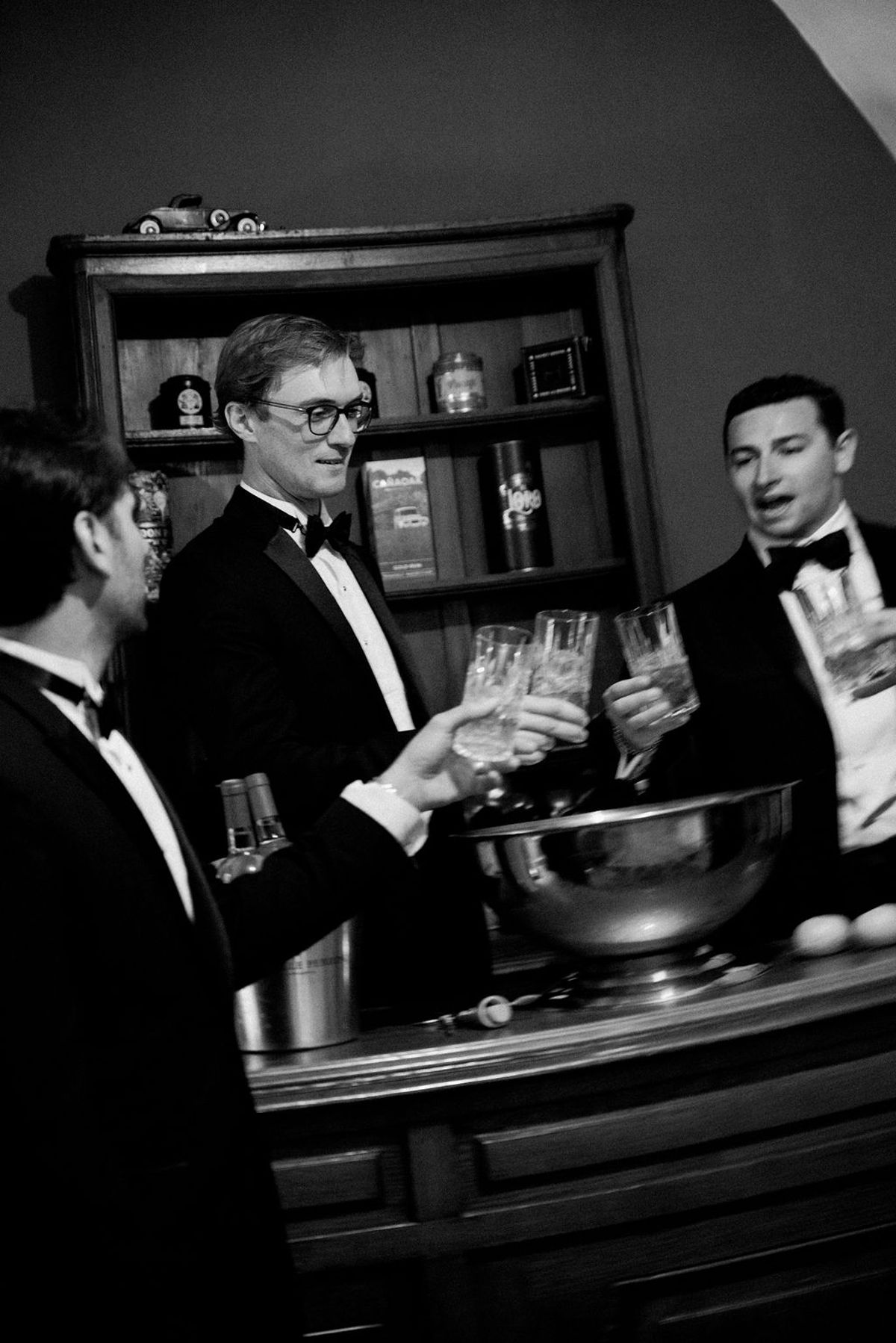 Three groomsmen in black tie raise crystal glasses in a toast at a wooden bar, in black and white.