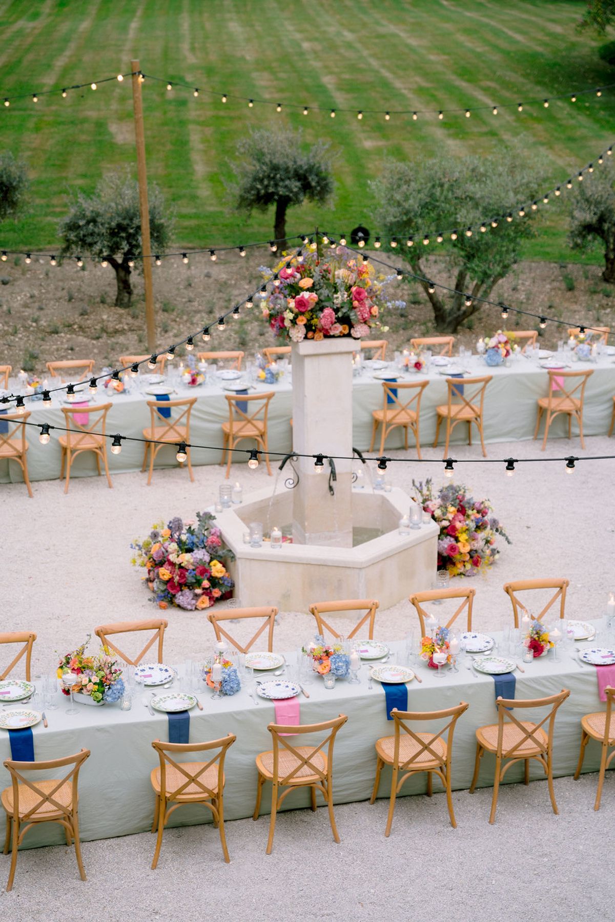 Long dining tables with wooden chairs and colourful flowers are arranged around a stone fountain on a lawn beneath strings of lights.