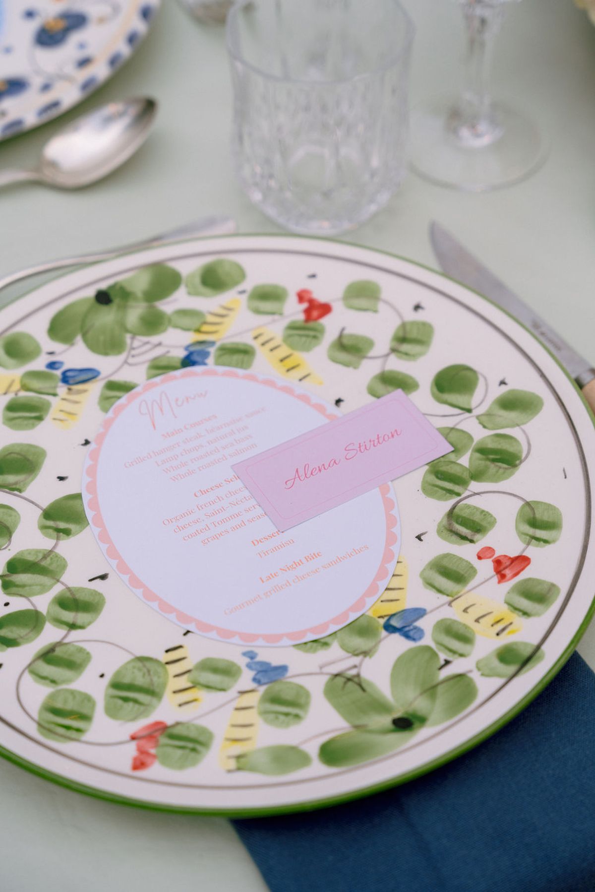 A hand-painted plate with a green leaf pattern holds a printed menu and a pink place card.