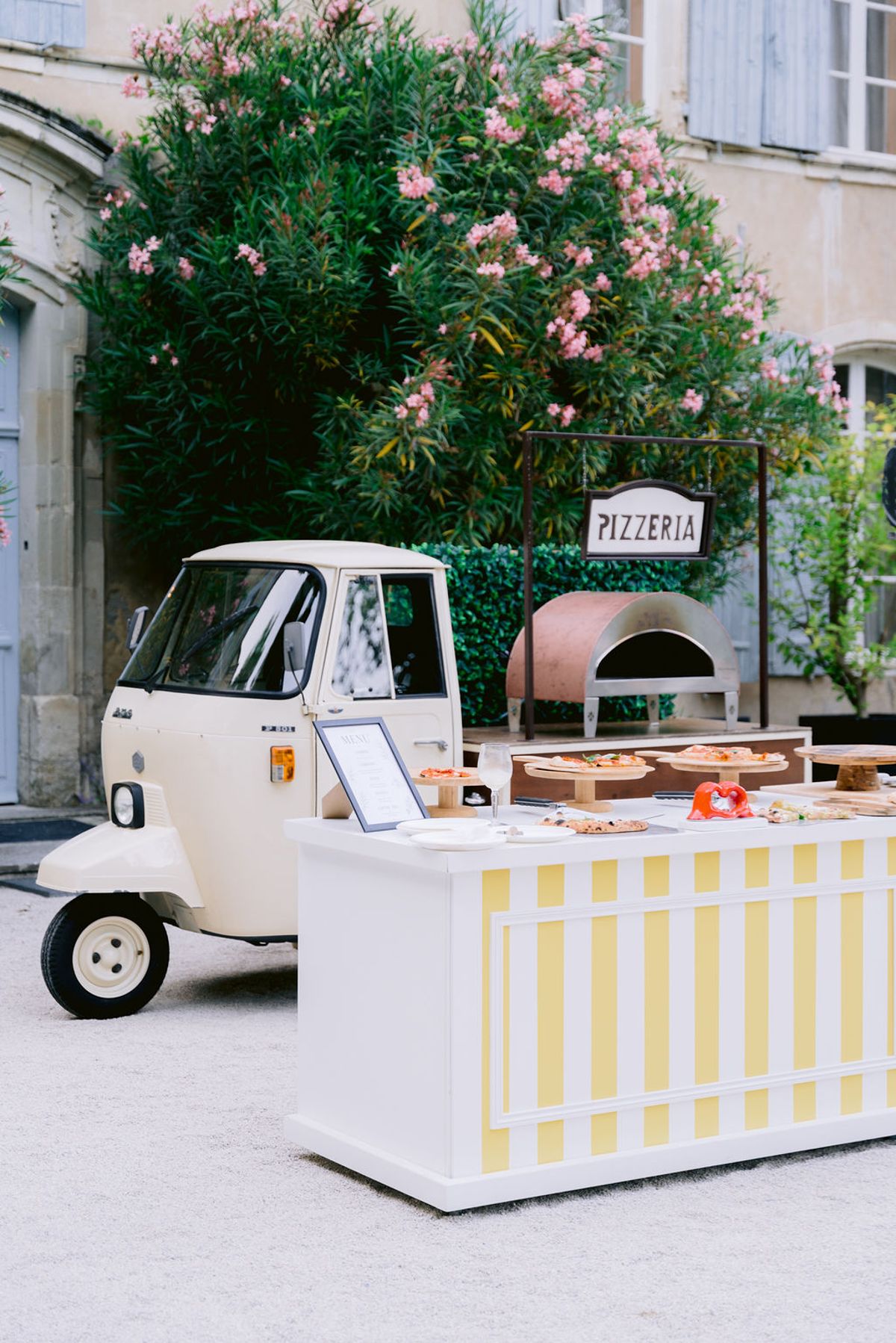 A small cream three-wheeled van with a pizza oven and a yellow-and-white striped counter stands before a stone building and pink flowering shrubs.