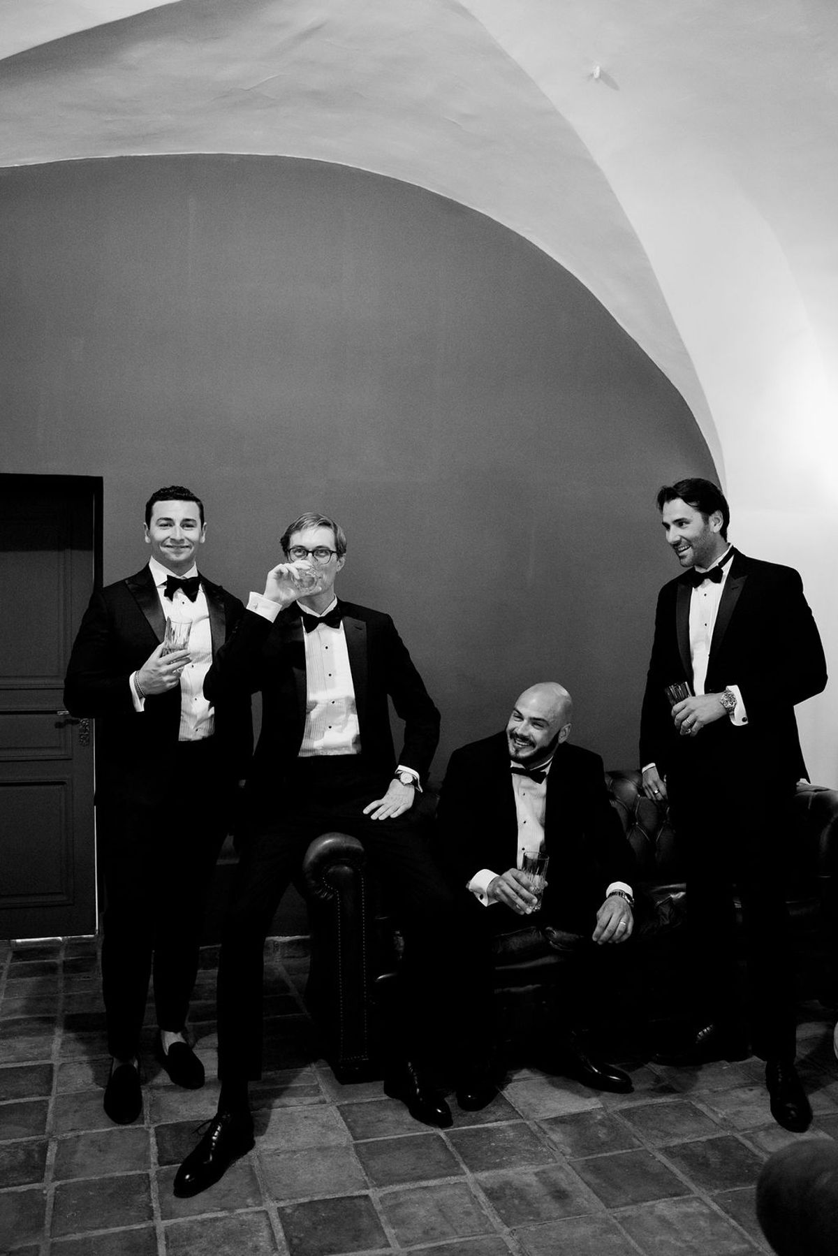 In black and white, four men in tuxedos stand and sit in a relaxed group beneath a curved stone ceiling.