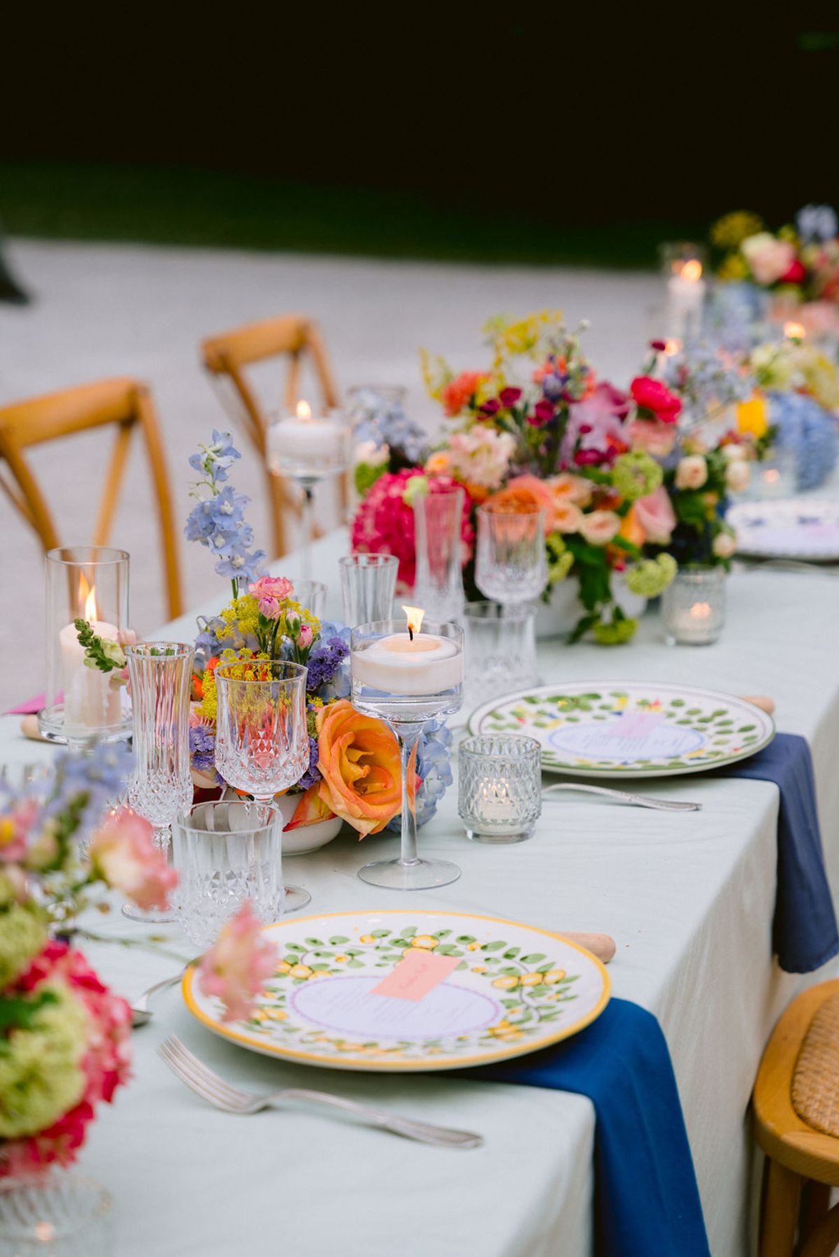 A long table dressed with a pale cloth, bright floral centrepieces, painted plates, crystal glasses and candles, set for a dinner outdoors.