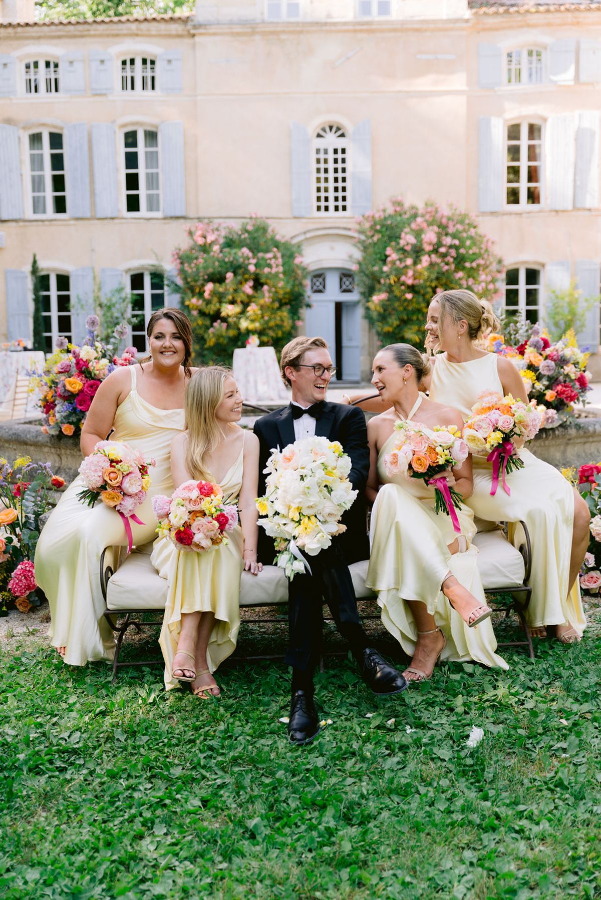 A groom holding a white bouquet sits on a bench between bridesmaids in pale yellow dresses with colourful bouquets, in front of a chateau.