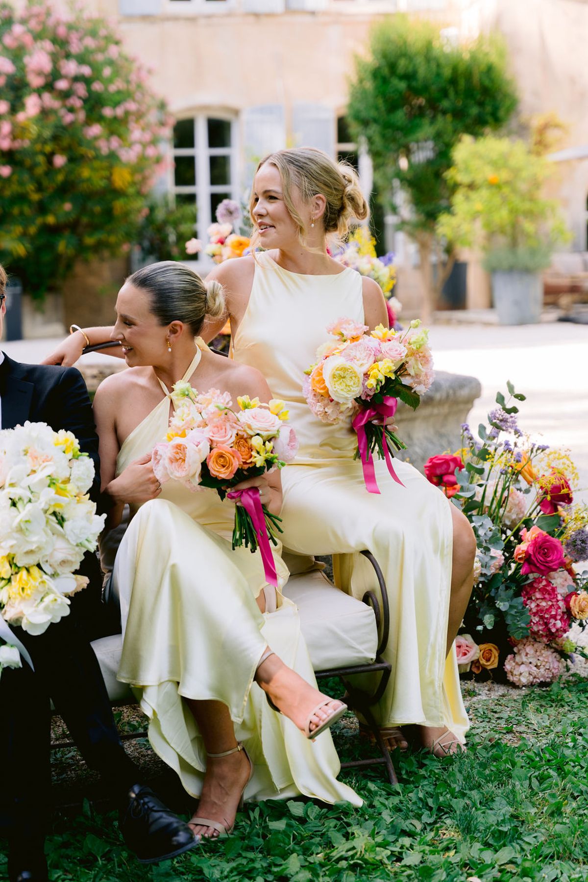 Two bridesmaids in pale yellow satin dresses sit holding pink and peach bouquets during the ceremony.