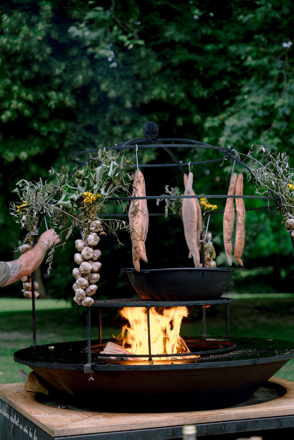 An open-fire grill hung with garlic, herbs and food over the flames, with a cook's hand reaching in.