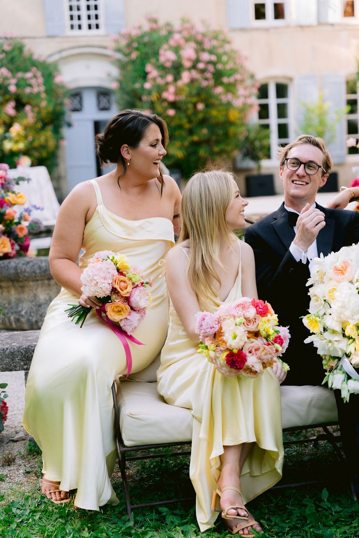 Two bridesmaids in pale yellow dresses holding colourful bouquets sit laughing on a cushioned bench beside a man in a dark suit.