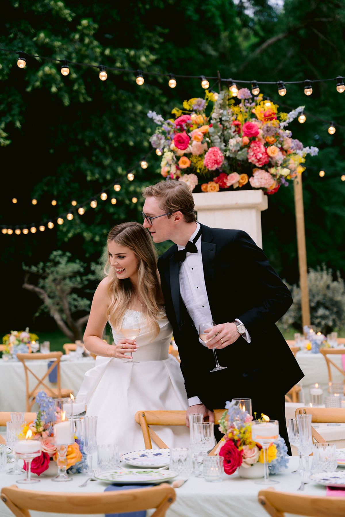 The bride and groom share a moment by the dinner tables, glasses in hand, under festoon lights and tall flowers.