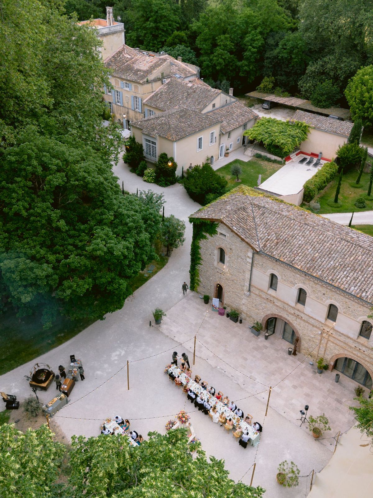 Seen from above, a stone courtyard surrounded by old buildings and trees holds a long dining table set for a meal.