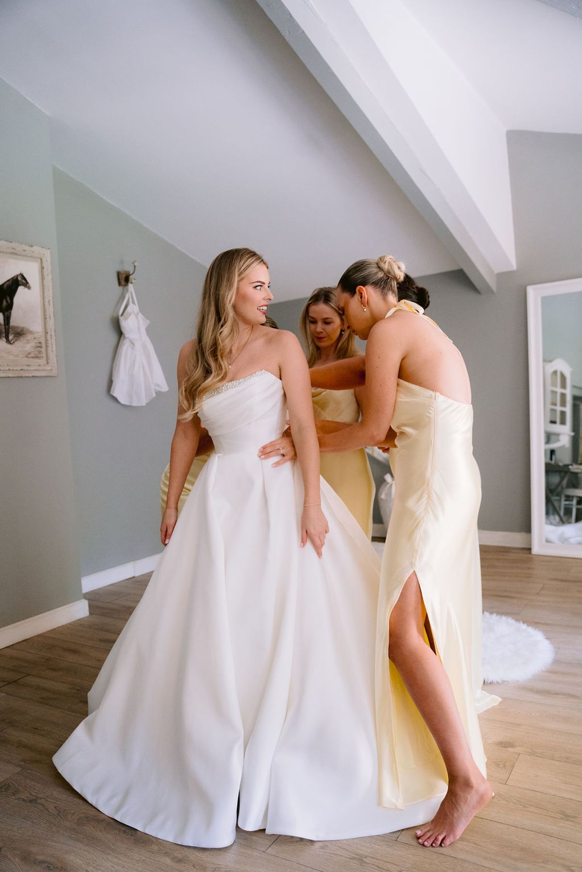 The bride in a strapless white gown stands in a pale room with two bridesmaids in yellow dresses, a dress hanging on the wall behind.