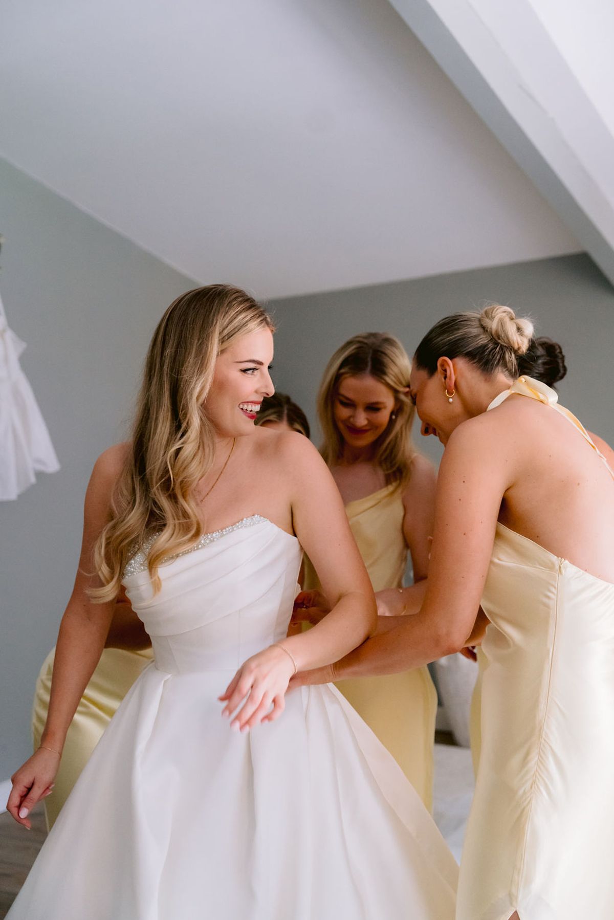 The bride in a strapless gown smiles as two bridesmaids in yellow dresses lean in and embrace her inside a light room.