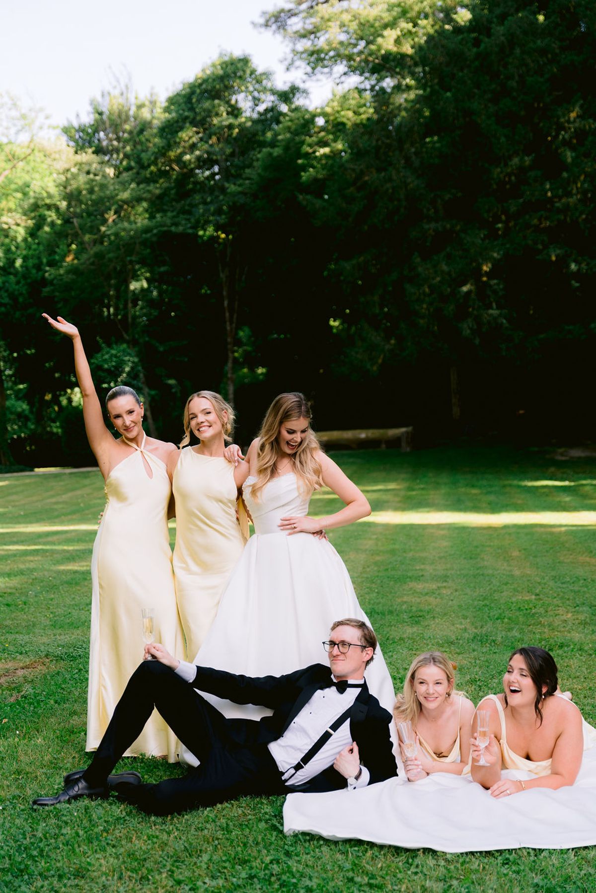 Bridesmaids in yellow strike playful poses on a lawn while a man in a tuxedo lies on the grass beside women laughing.