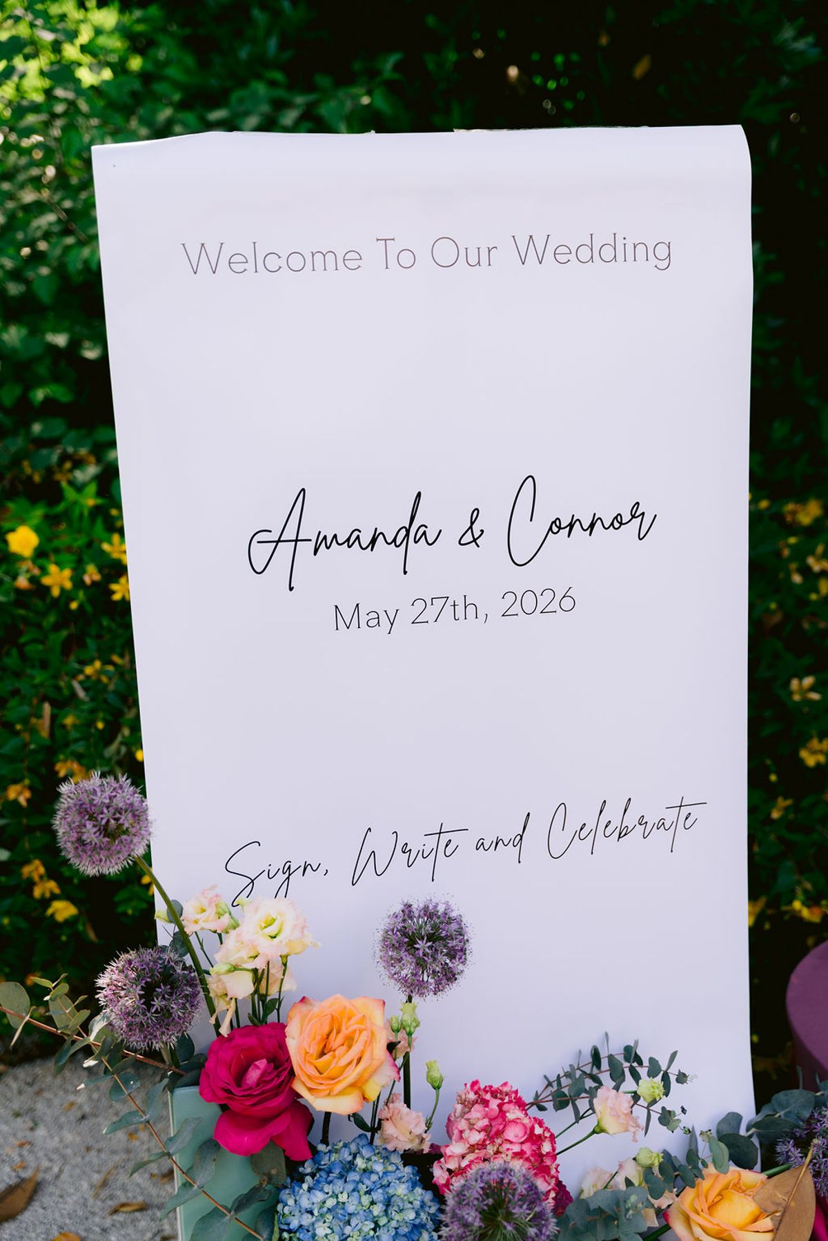 A tall white welcome sign with black script lettering stands among greenery, with colourful flowers arranged at its base.