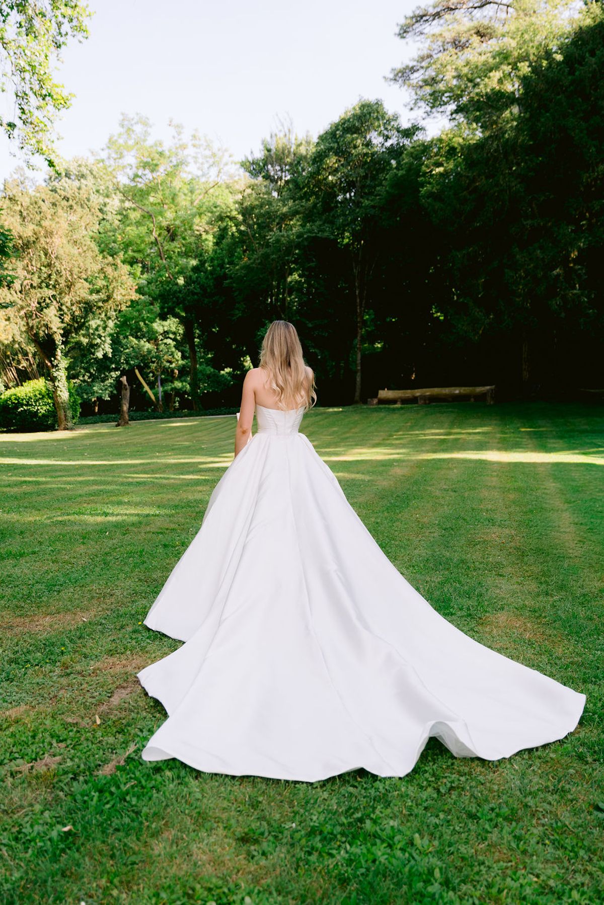 The bride, seen from behind in a white gown with a long train, walks across a green lawn towards tall trees.
