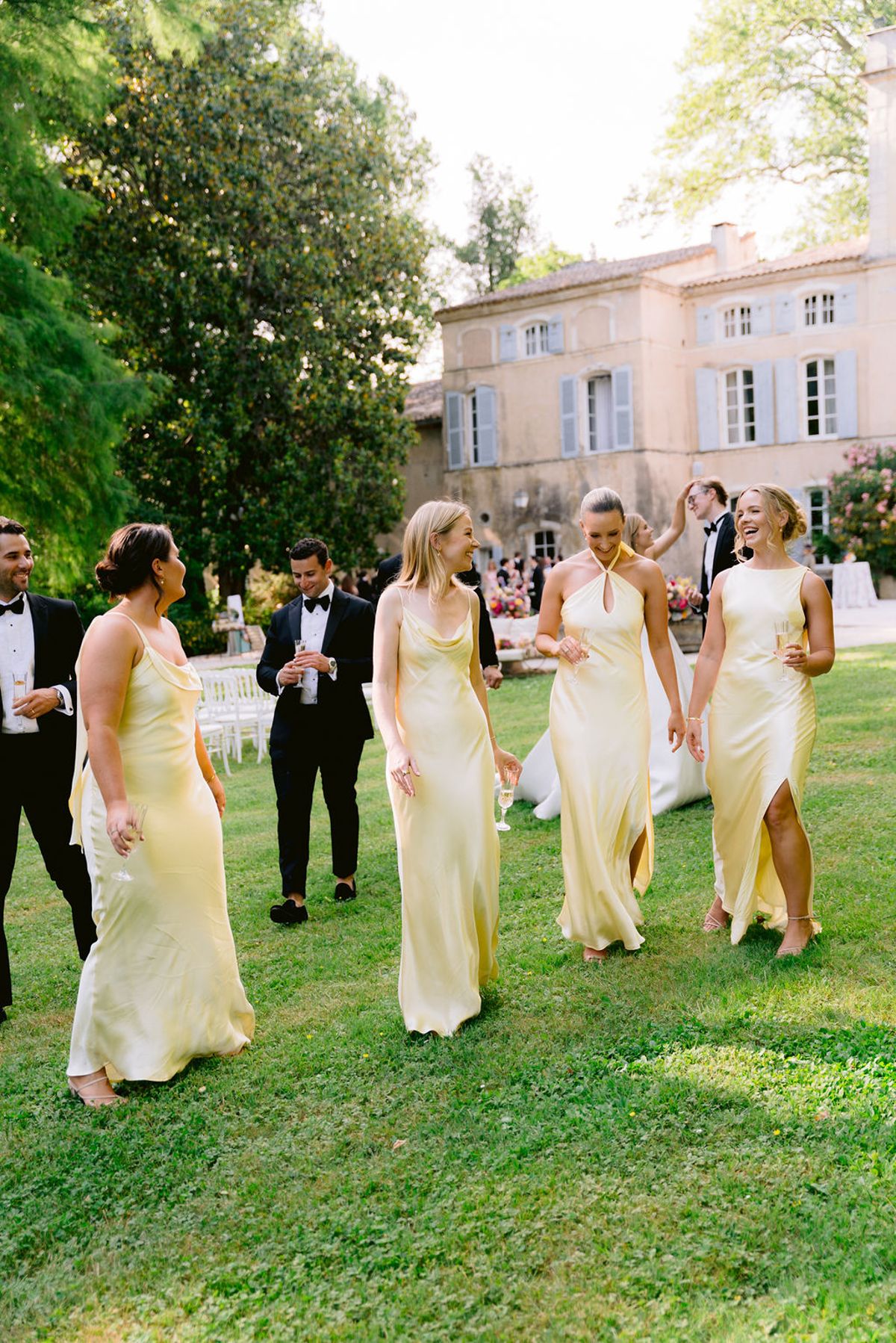 Bridesmaids in pale yellow satin dresses walk across the lawn with groomsmen in black tie behind them.