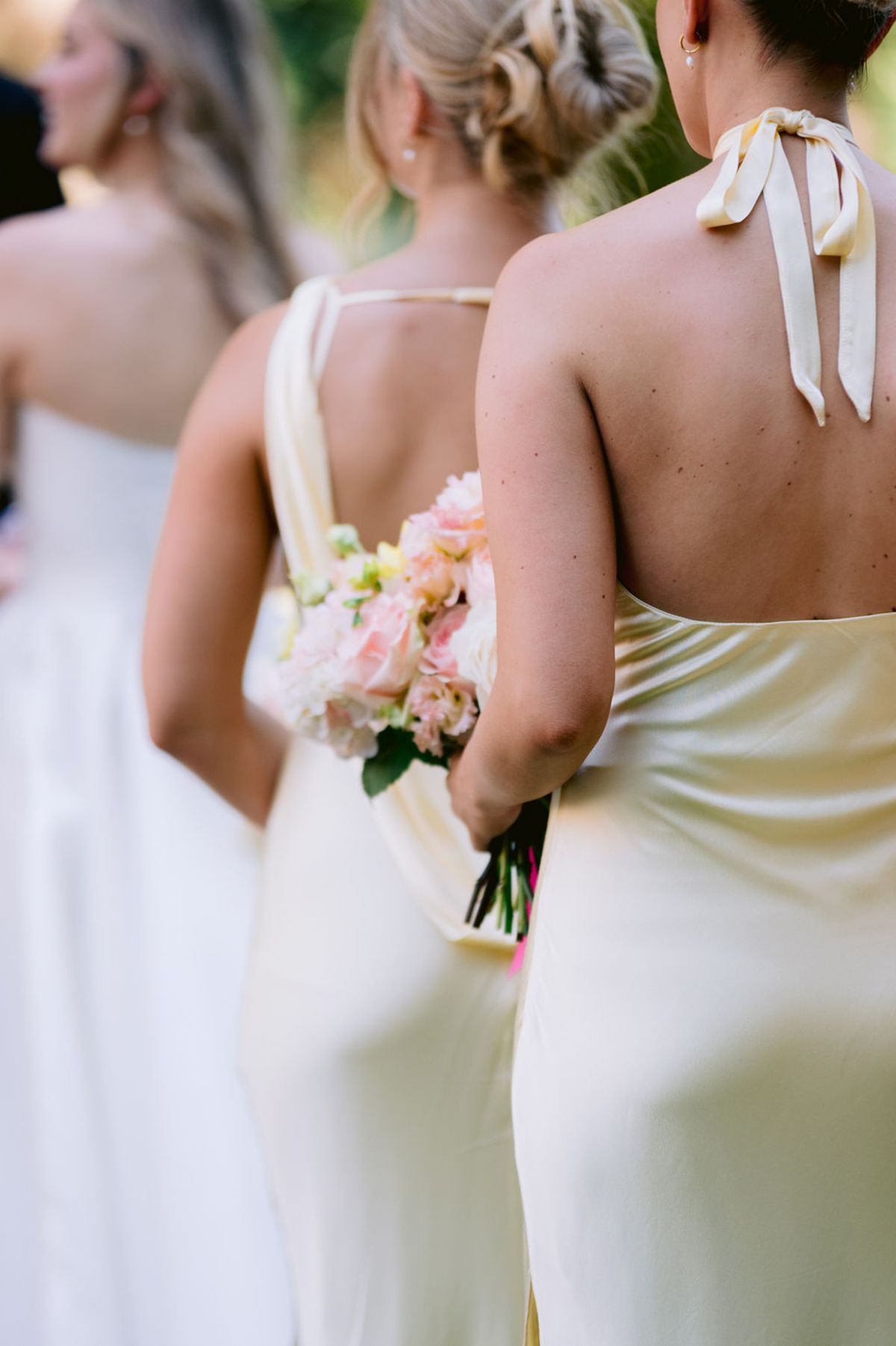 Seen from behind, bridesmaids in pale yellow satin dresses stand together, one holding a pink bouquet.