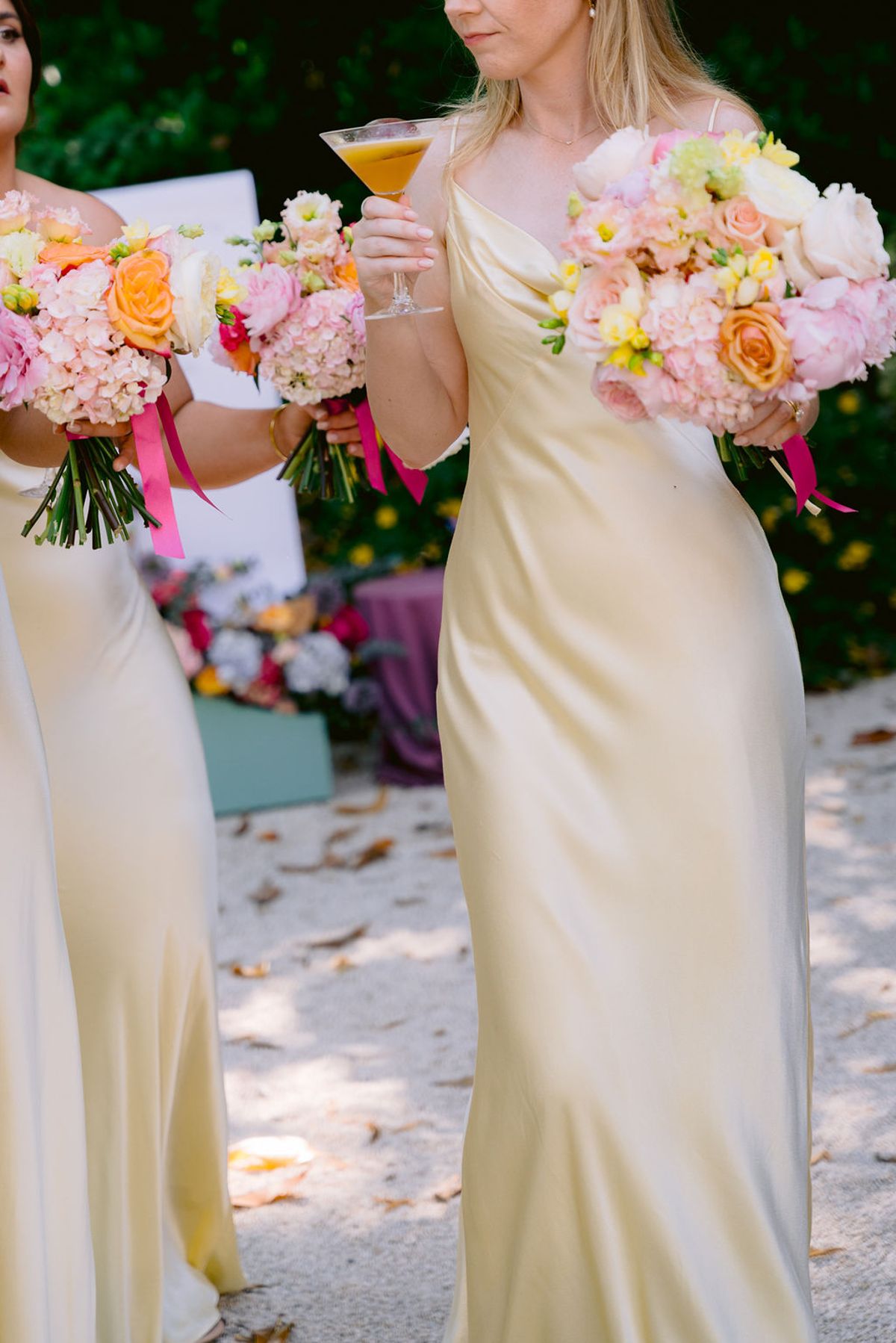 Bridesmaids in yellow satin dresses hold bright pink and orange bouquets, one also holding an orange cocktail, on a gravel path.
