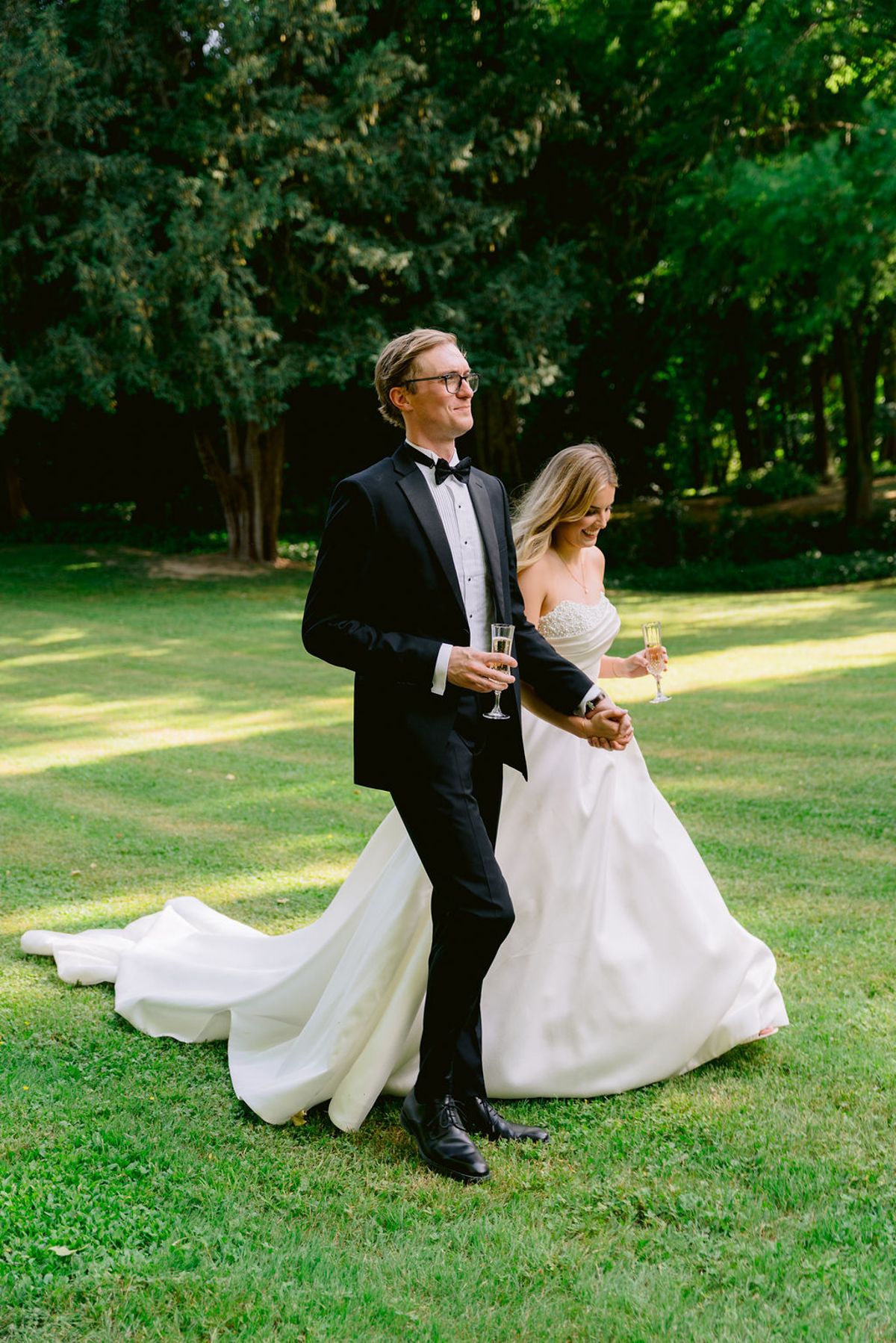 The groom in a tuxedo holds a glass and walks across a lawn beside the bride, her long train trailing behind, with trees beyond.