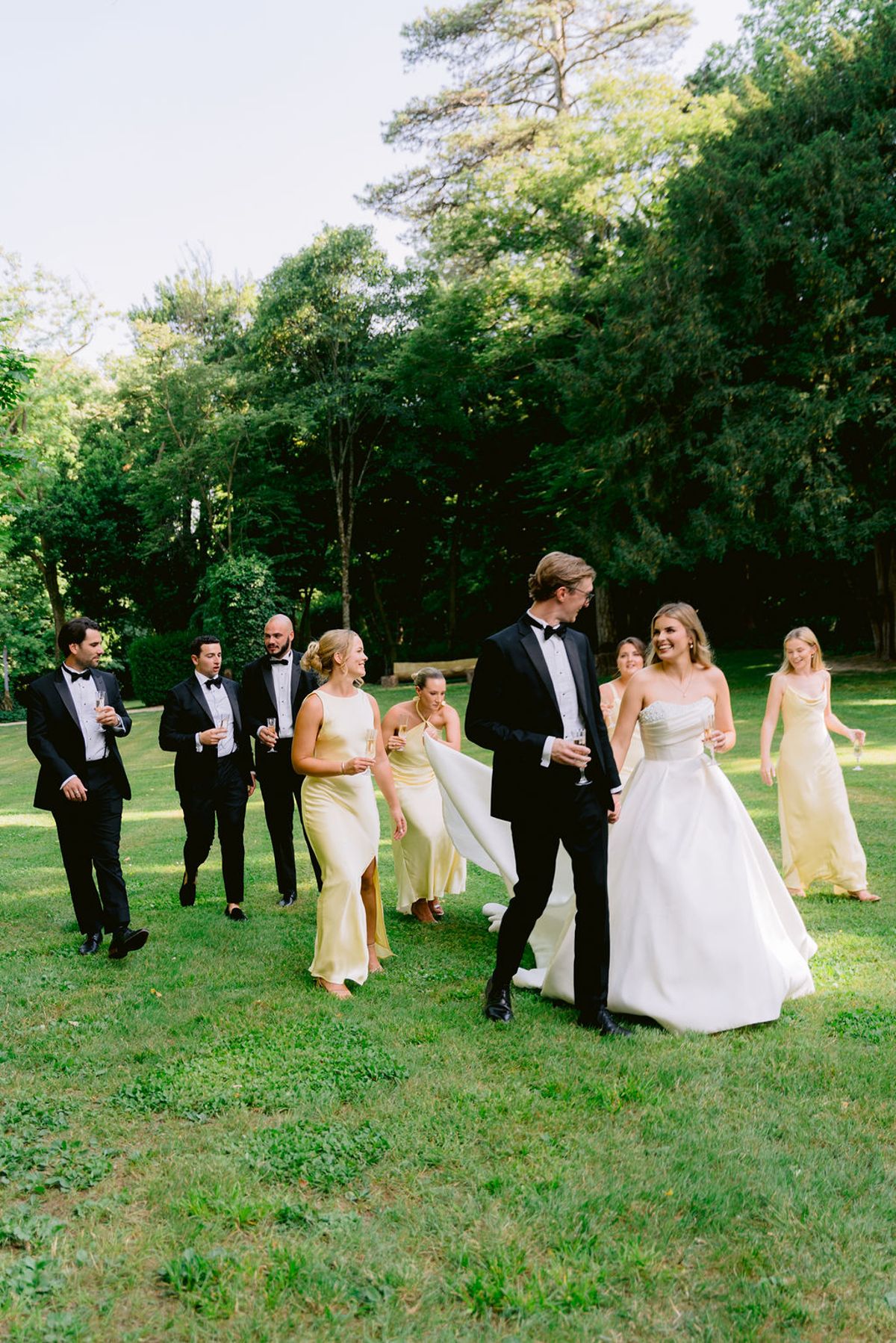 The couple walk across a sunlit lawn beside bridesmaids in pale yellow dresses and groomsmen in black tuxedos, with tall trees behind.