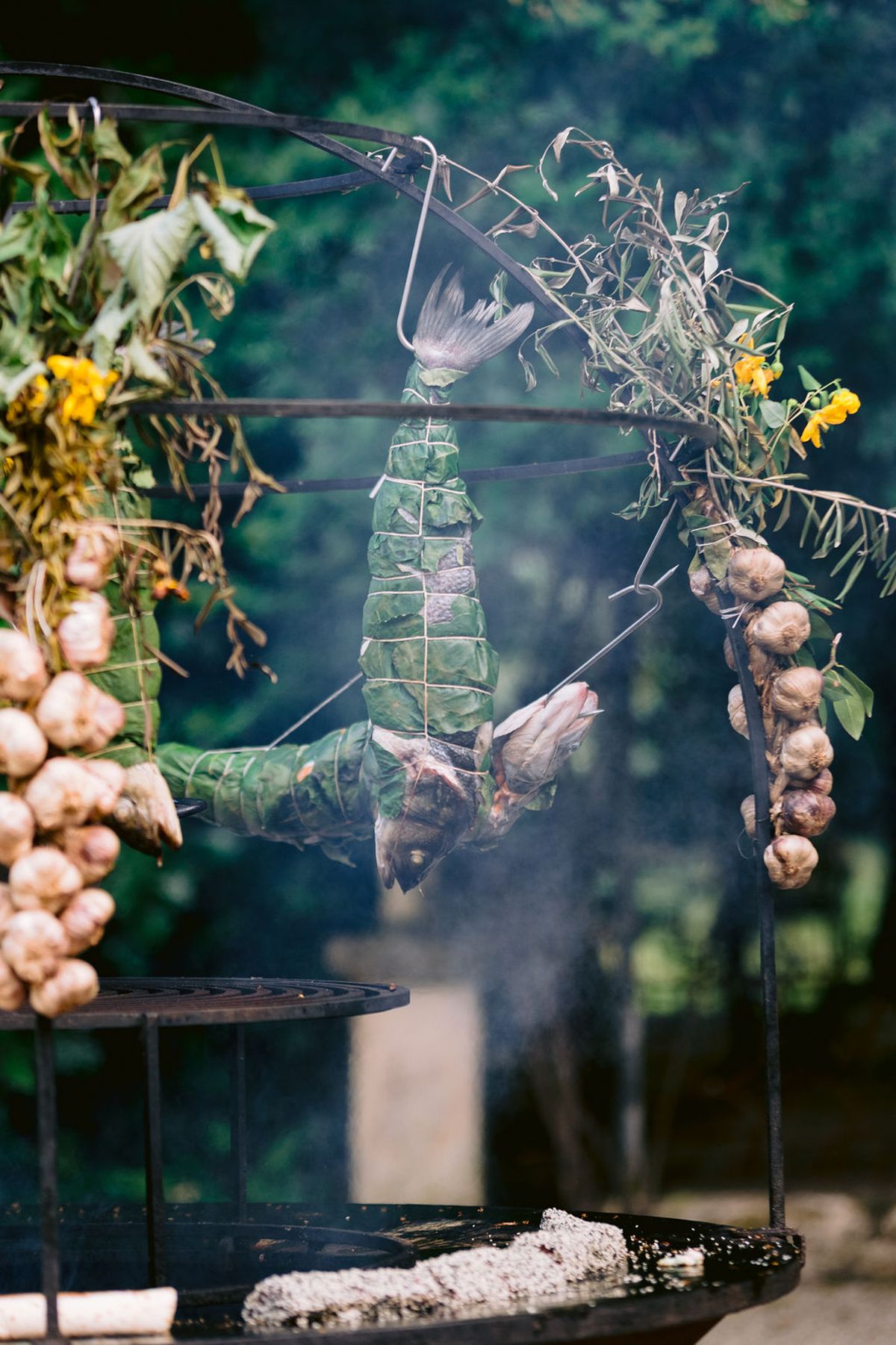 A food parcel hangs upside down over a smoking fire pit beneath a metal arch decorated with garlic bulbs and flowers.
