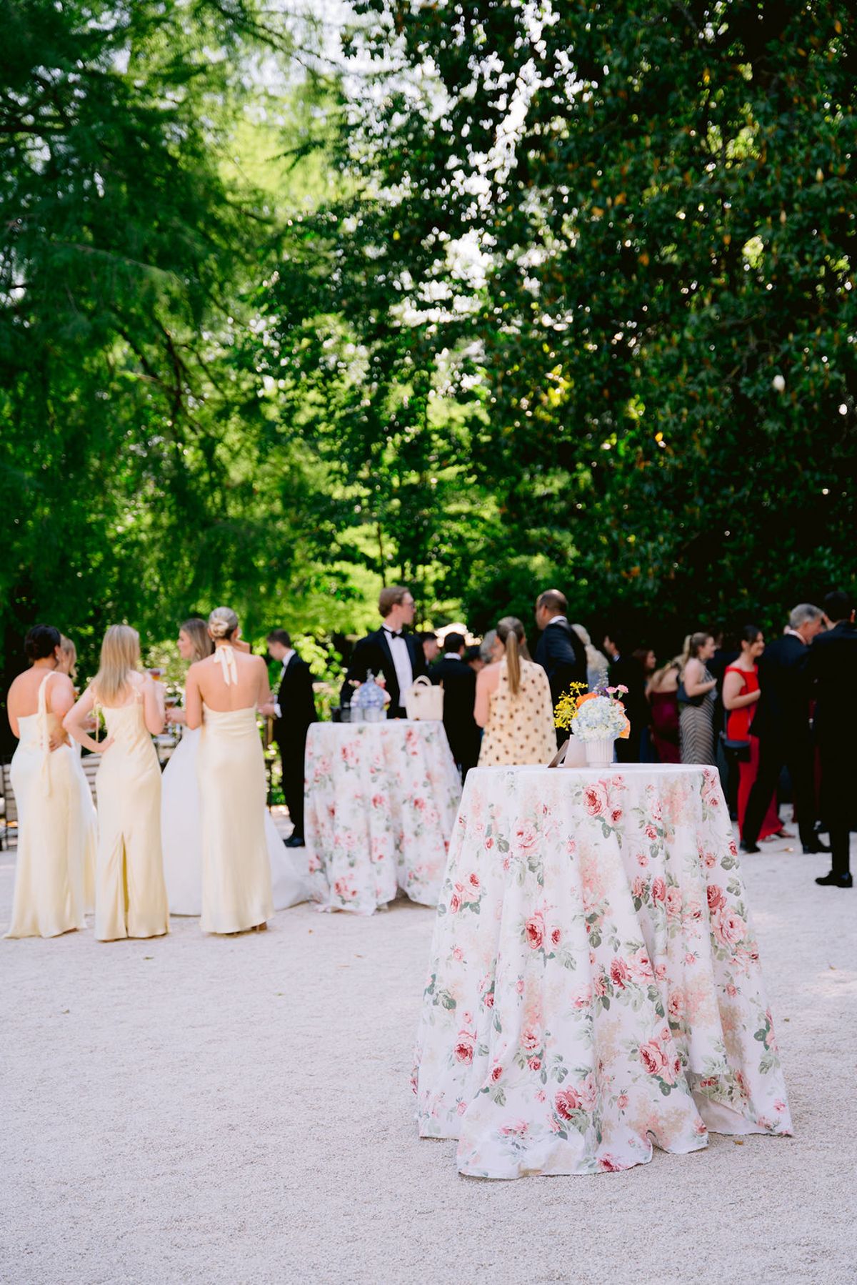 Guests in formal wear and yellow dresses stand around tall tables with floral cloths on a gravel area beneath tall green trees.
