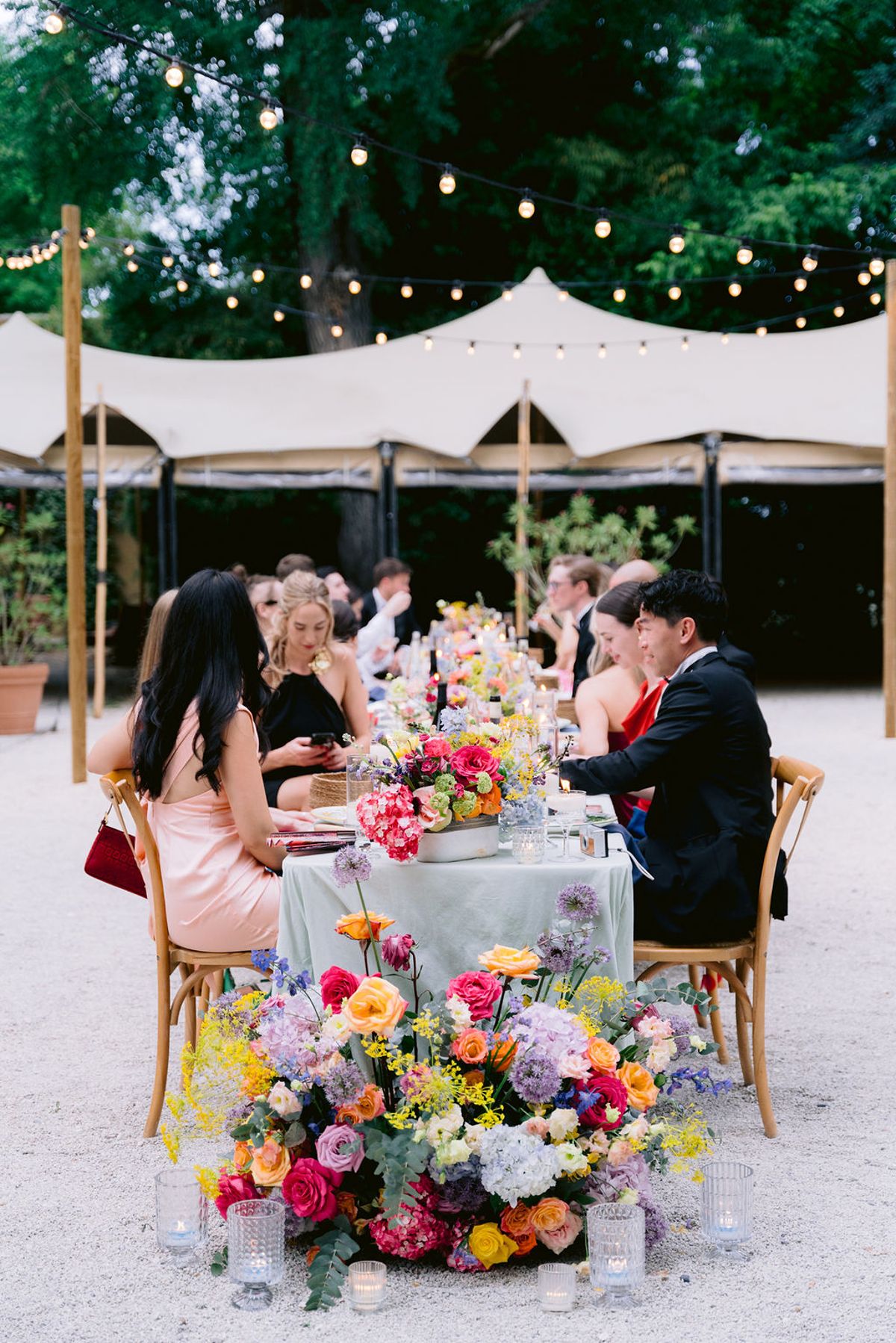 Guests dine at a table with a colourful flower centrepiece beneath a white marquee and string lights, with large floral arrangements in front.