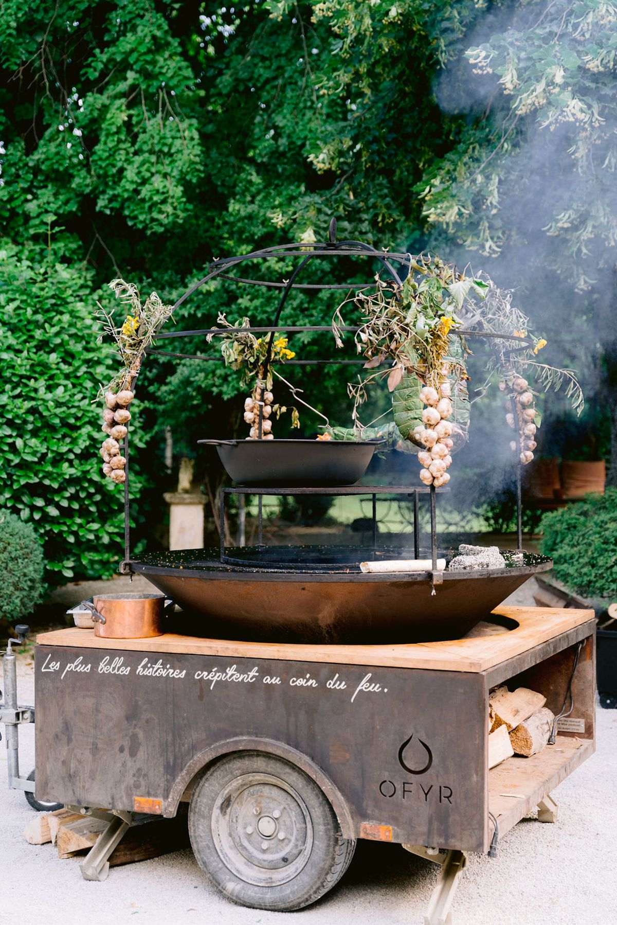 A cast iron fire bowl on a wooden trailer sends up smoke beneath a metal arch hung with garlic bulbs and flowers.