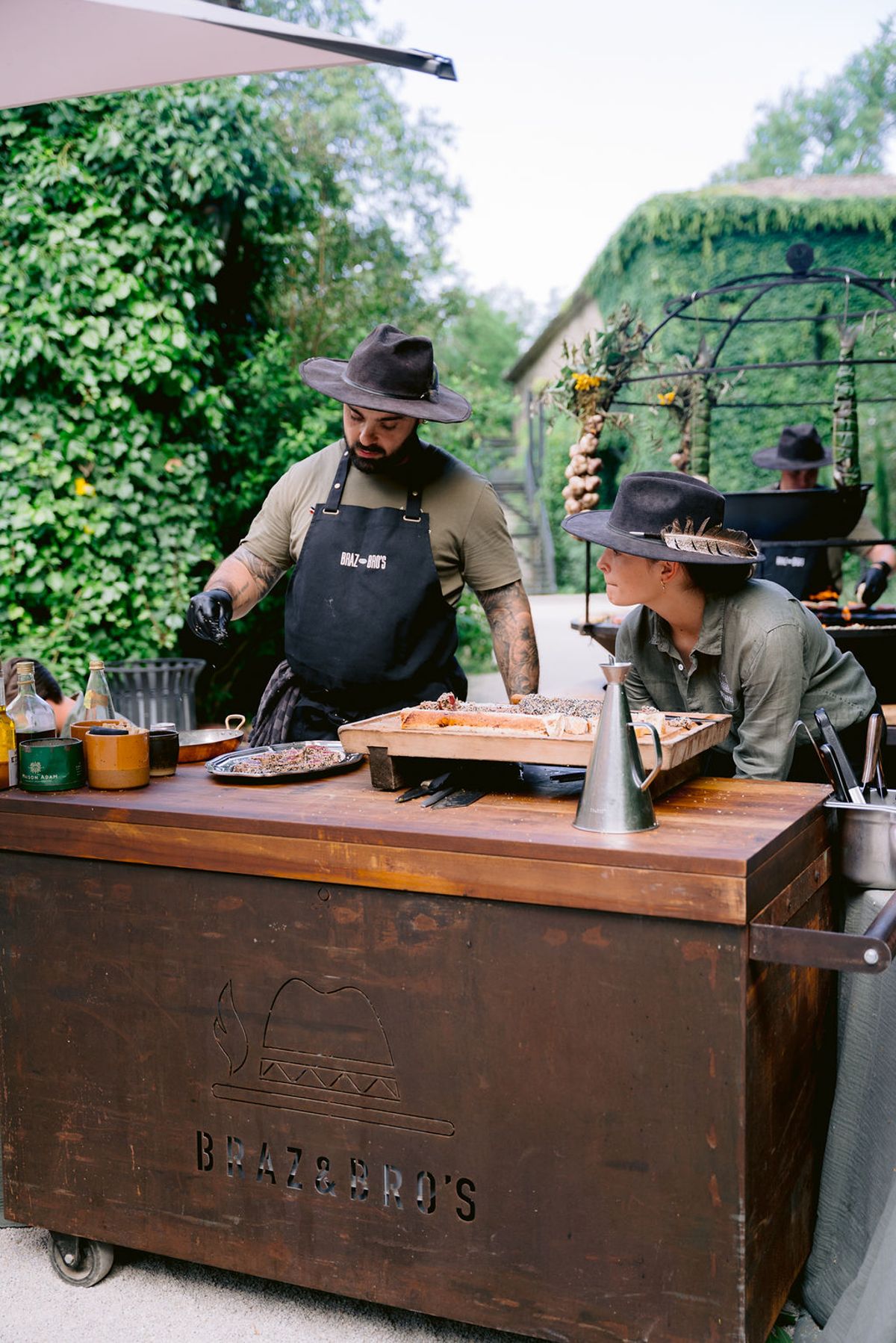 Two cooks in wide-brimmed hats prepare food at a wooden open-air kitchen cart in the garden.