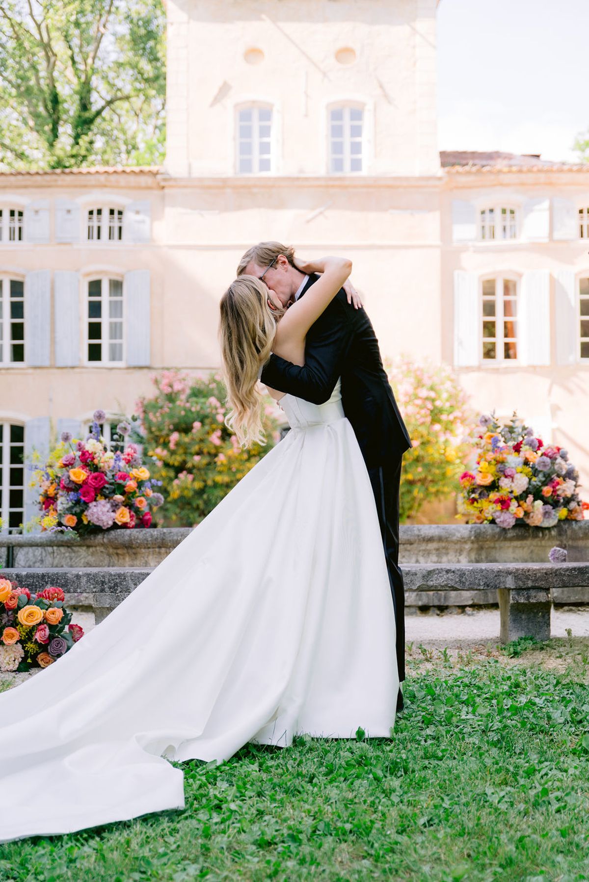 The couple embrace and kiss in front of a pale stone chateau, the bride's long train spread across the grass beside bright flowers.
