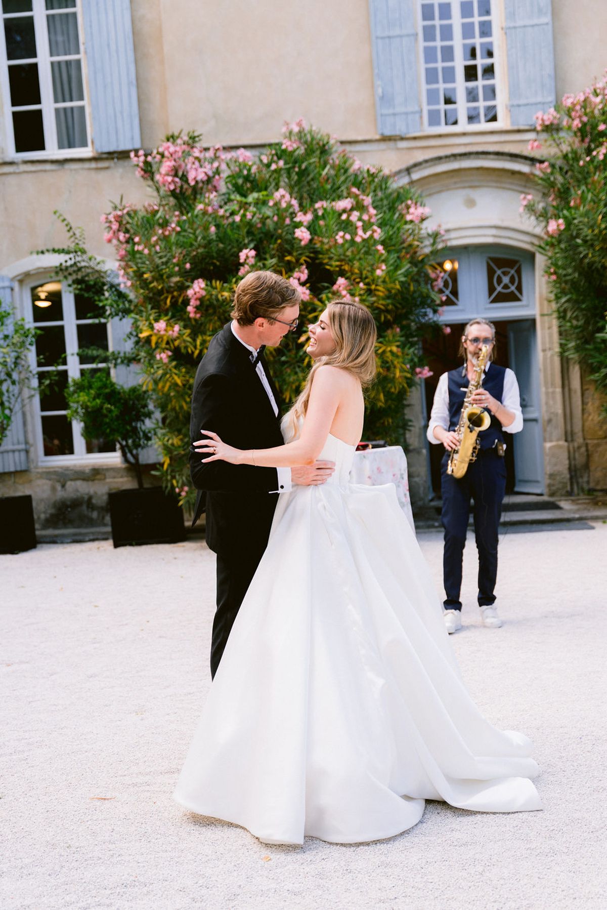 The bride in a white gown and the groom in black embrace on gravel in front of a chateau as a saxophonist plays behind them.