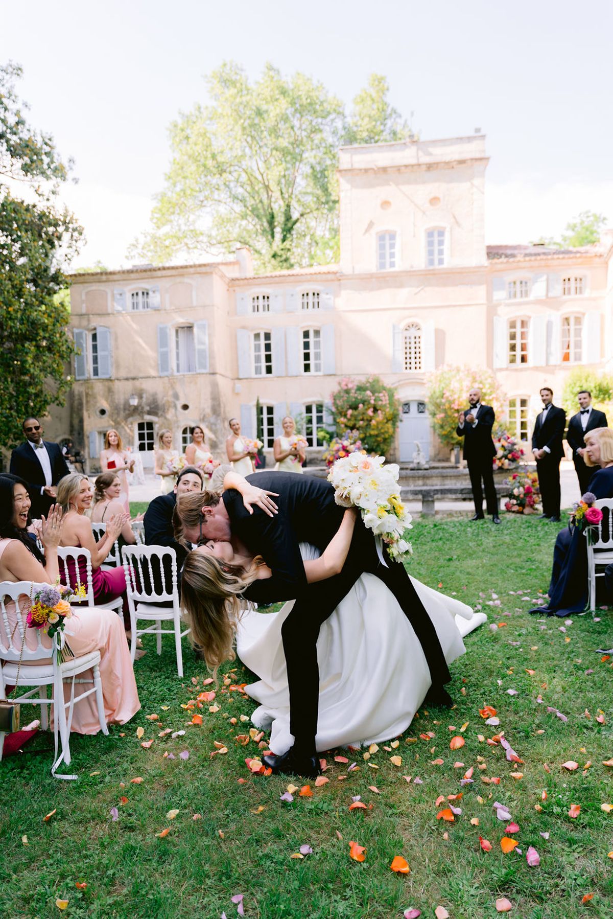 The groom dips the bride for a kiss on a petal-strewn aisle between rows of seated guests, with the chateau behind.