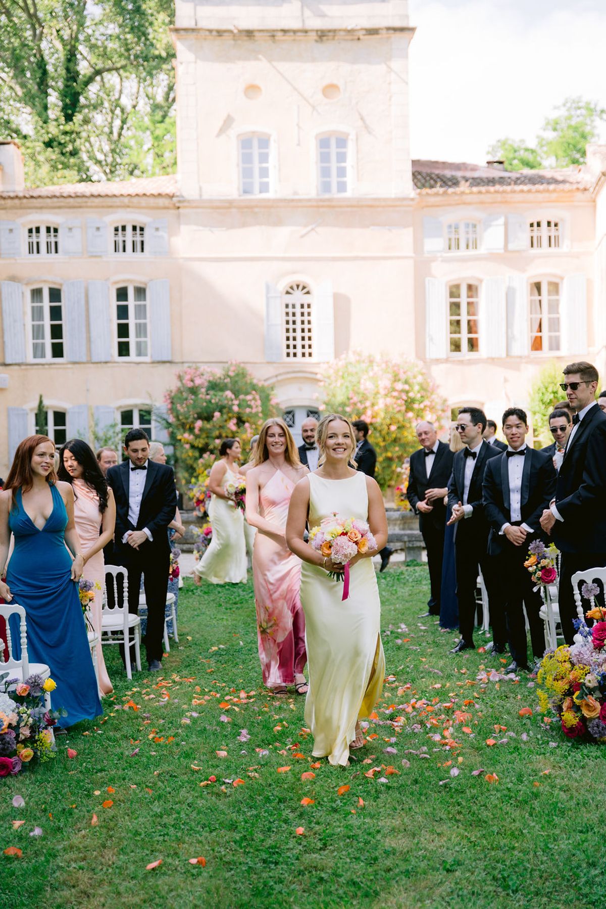 A bridesmaid in a pale dress carries a bouquet down an aisle scattered with petals, between standing guests, with the chateau behind.