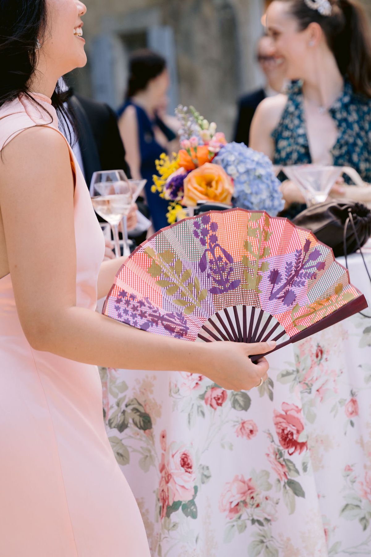 A guest holds a printed pink and purple fan beside a cocktail table with flowers and glasses.