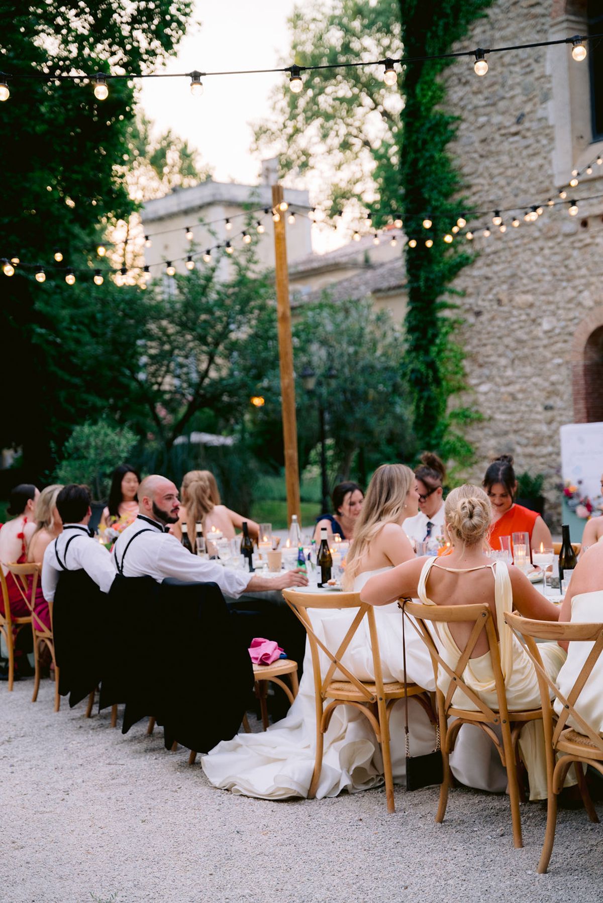 Guests sit at a long table on a gravel courtyard beneath string lights, with a stone building and climbing ivy behind.