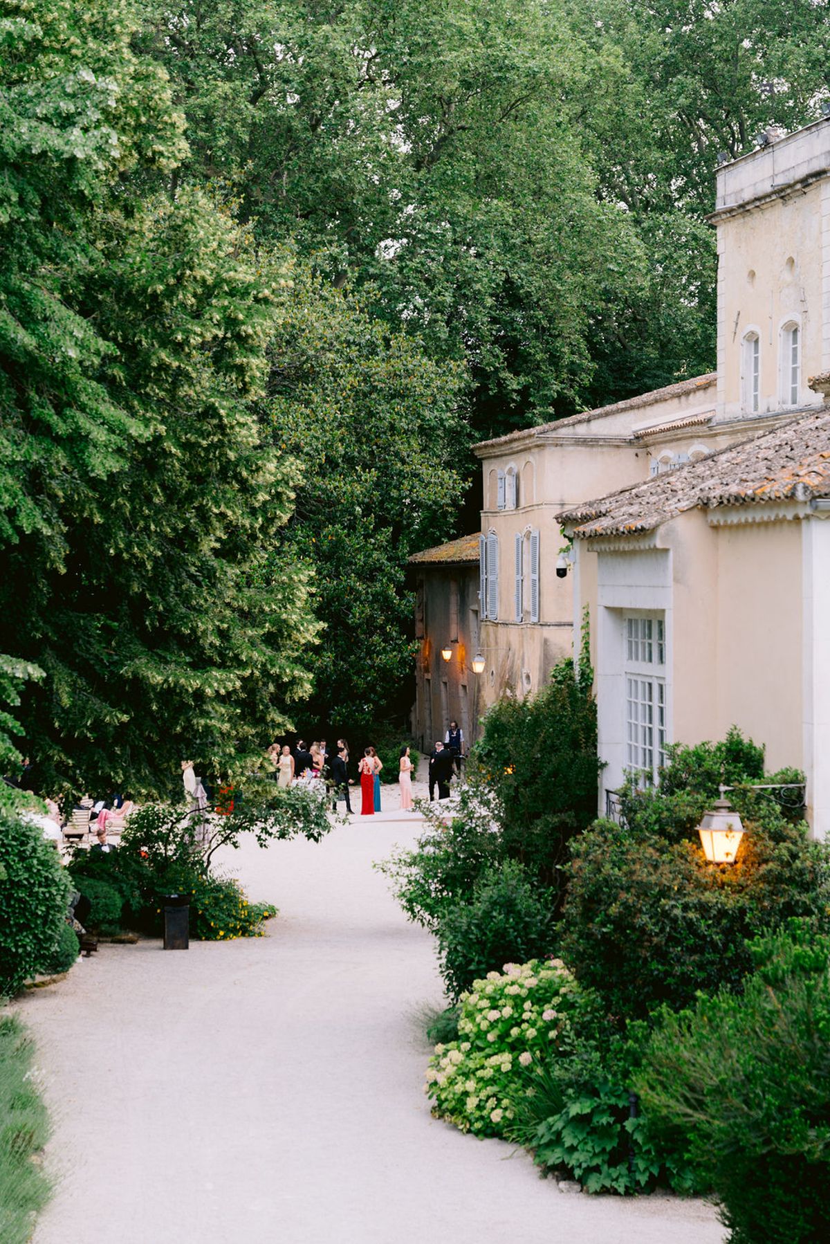 Guests gather at the end of a gravel path between tall trees and the pale stone buildings of the château.