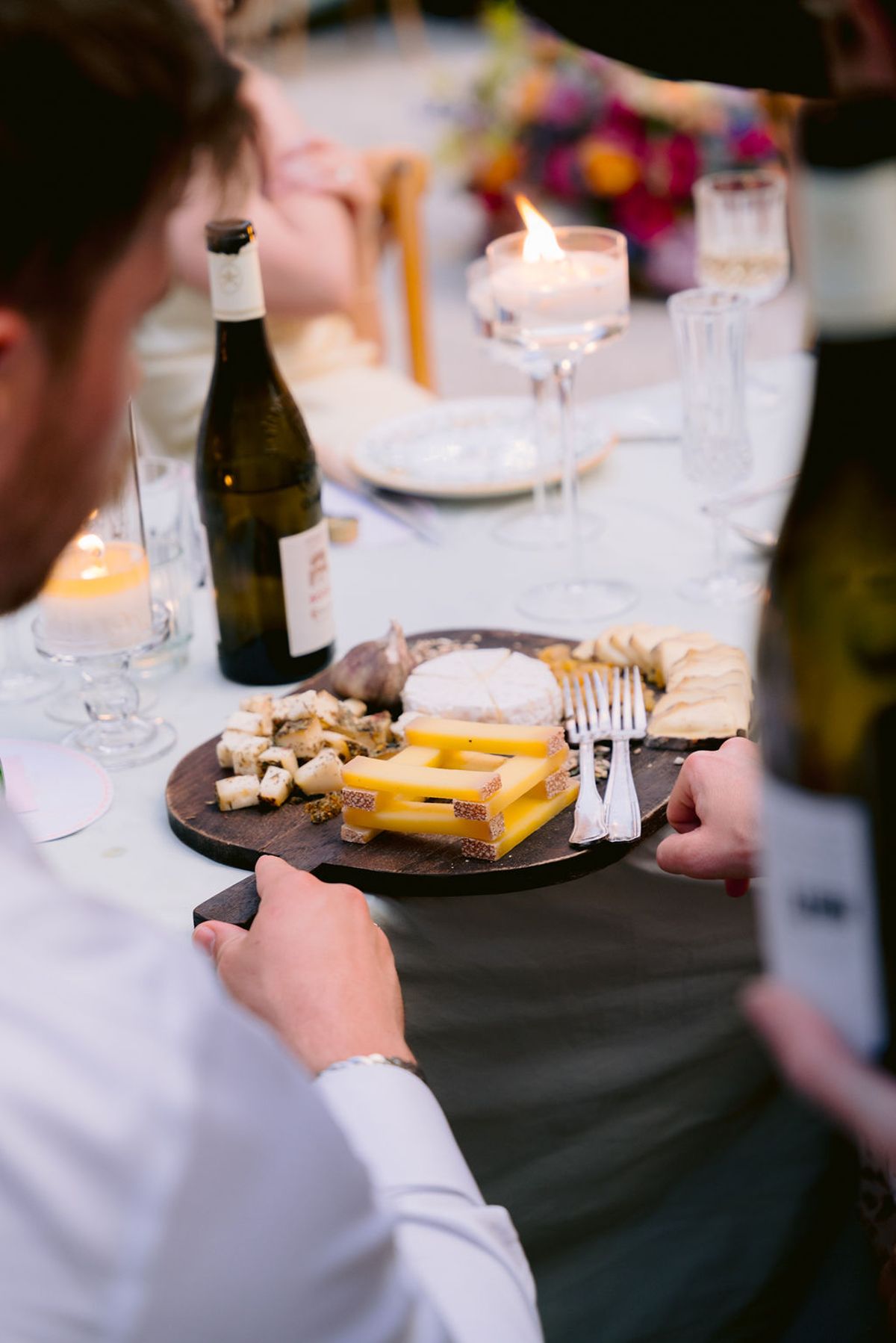 A guest holds a wooden board of cheeses beside candles, wine and glasses on the dinner table.