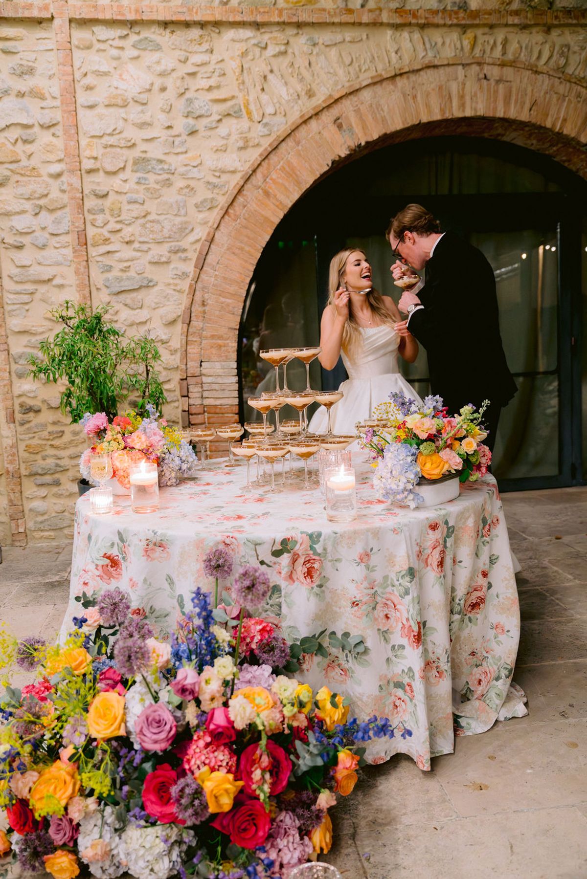 The couple share a kiss at a round table covered in a floral cloth, with bright bouquets and a stone arch behind.