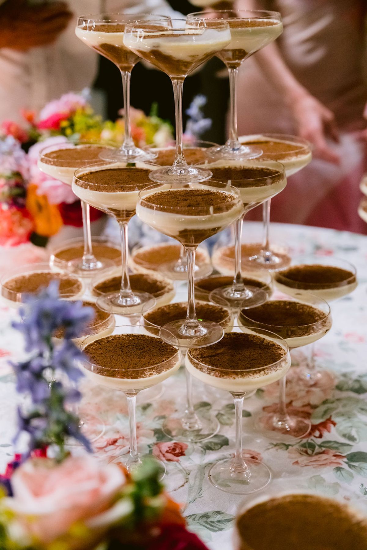 A stacked pyramid of coupe glasses holds creamy brown cocktails on a floral tablecloth, with bright flowers in the foreground.