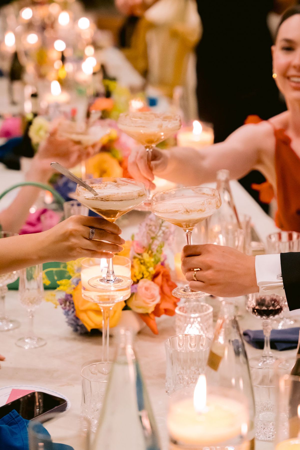 Guests reach across a candlelit table to clink coupe glasses among flowers, with one smiling woman in the background.