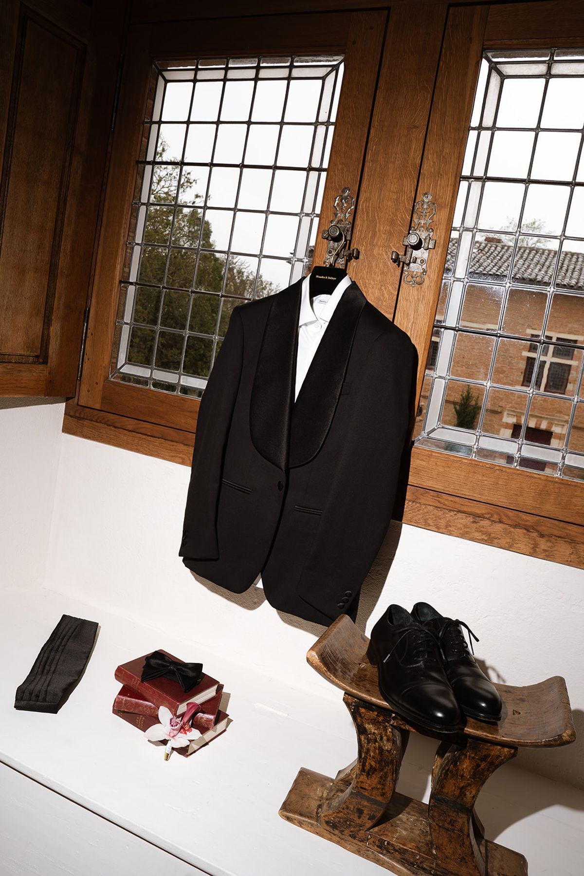 Groom tuxedo hanging by a leaded window, with shoes on a wooden stool and a bow tie on old books