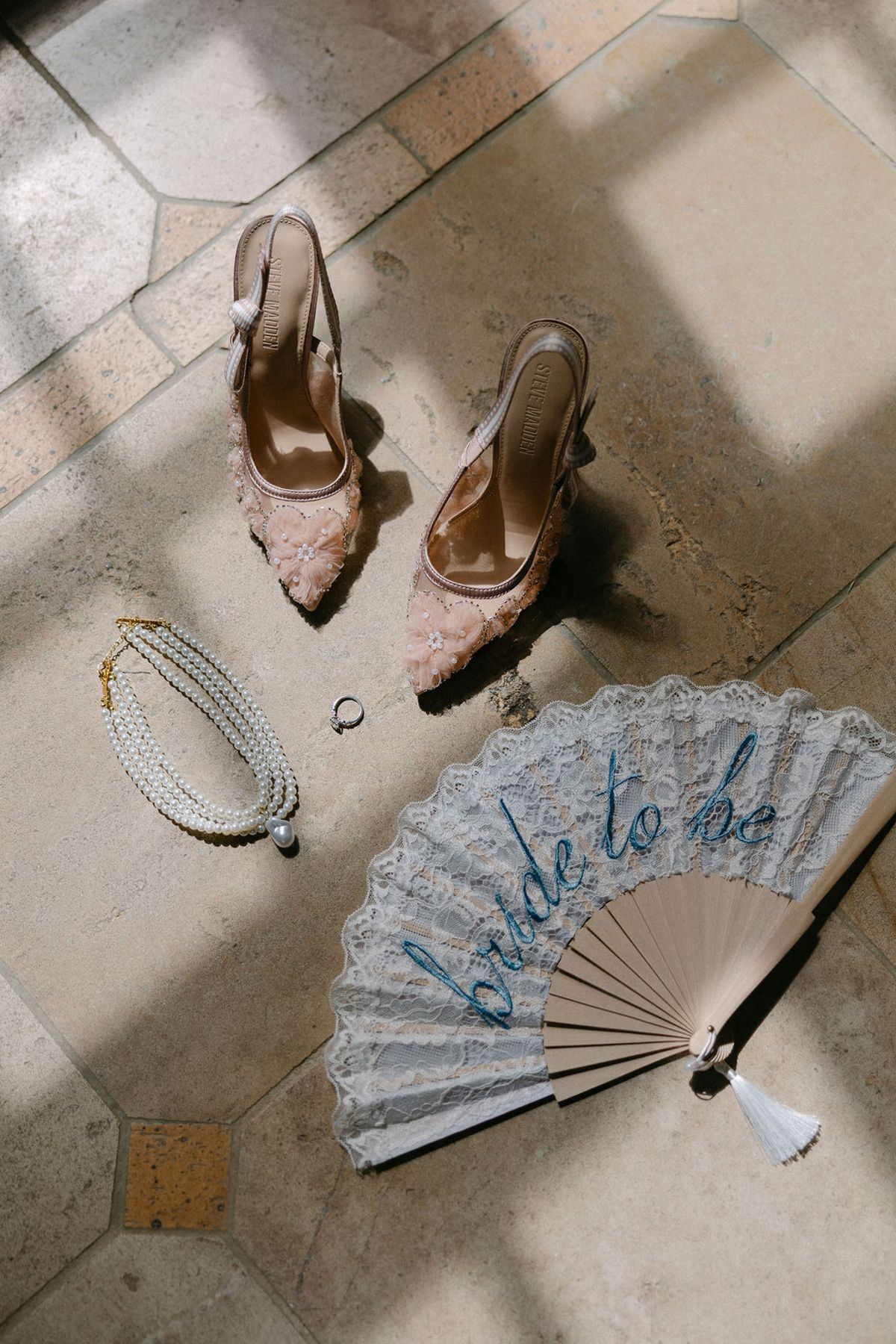 Flat lay of the bride's shoes, jewelry and a fan reading 'bride to be' on stone steps.