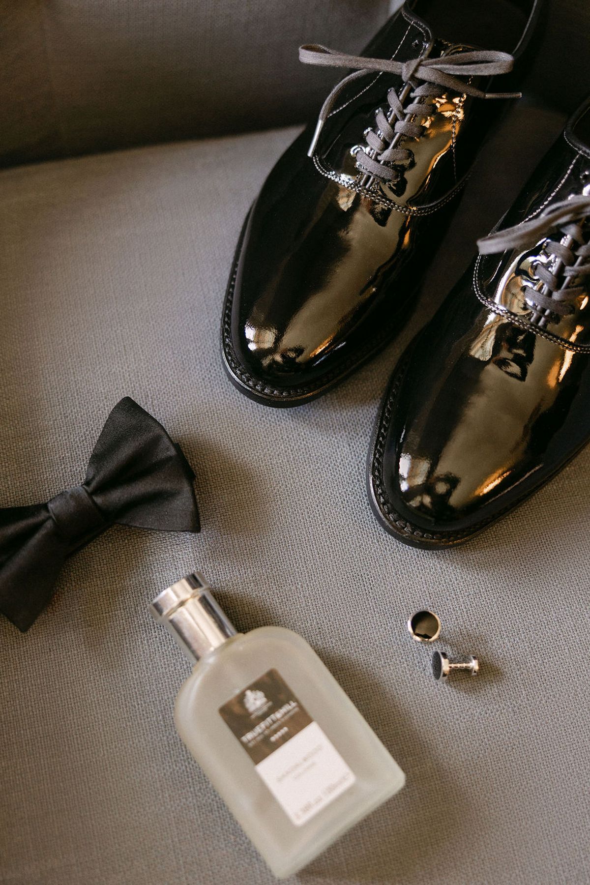 Flat lay of the groom's bow tie, patent shoes and cologne on linen.