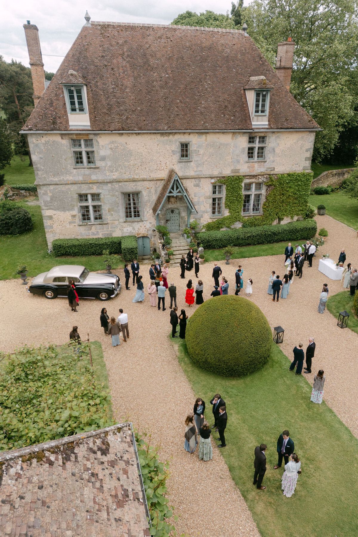 Aerial view of guests gathered on the gravel drive during the cocktail hour, vintage car parked alongside.