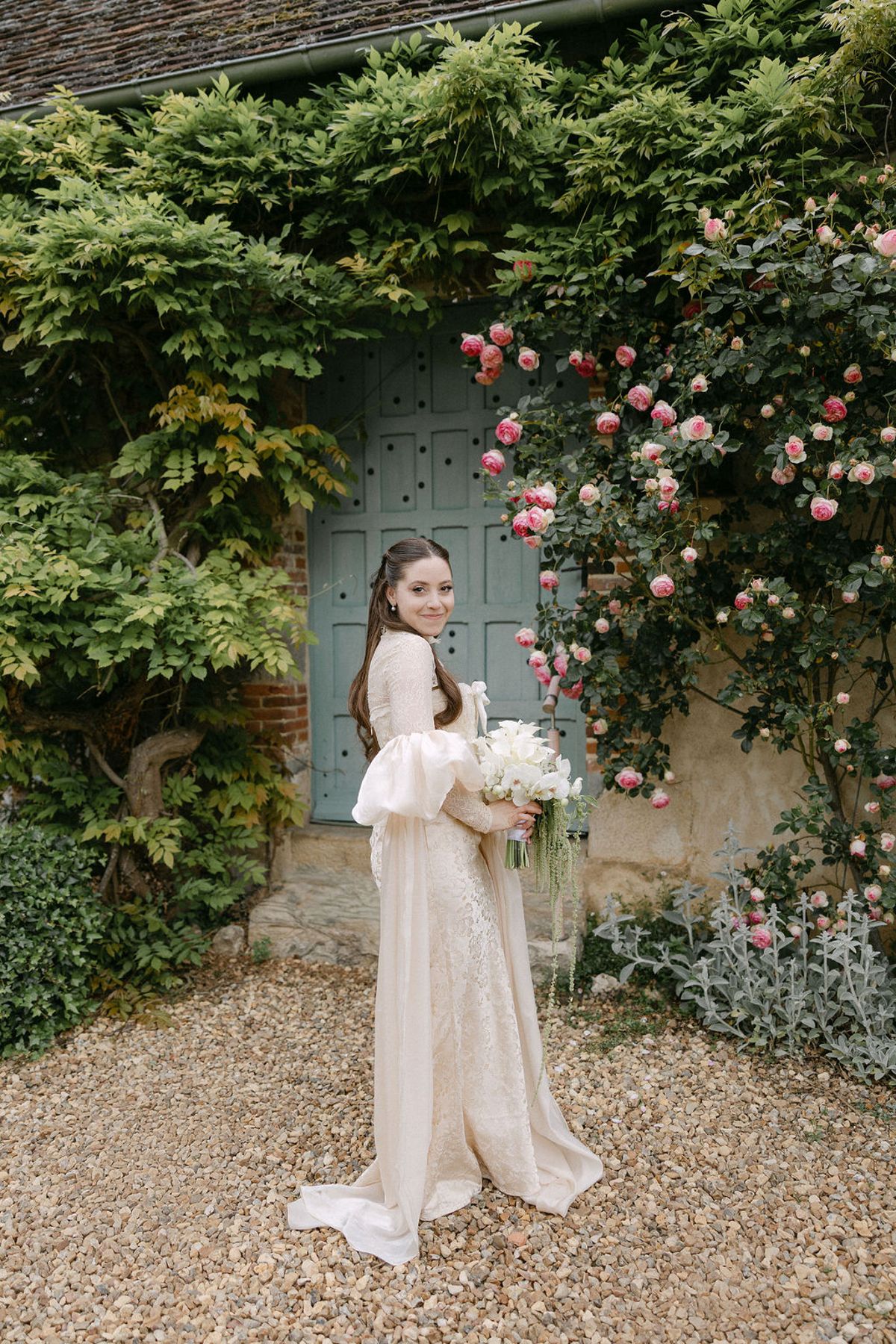 Olivia alone beside the rose-covered blue door, holding her bouquet.