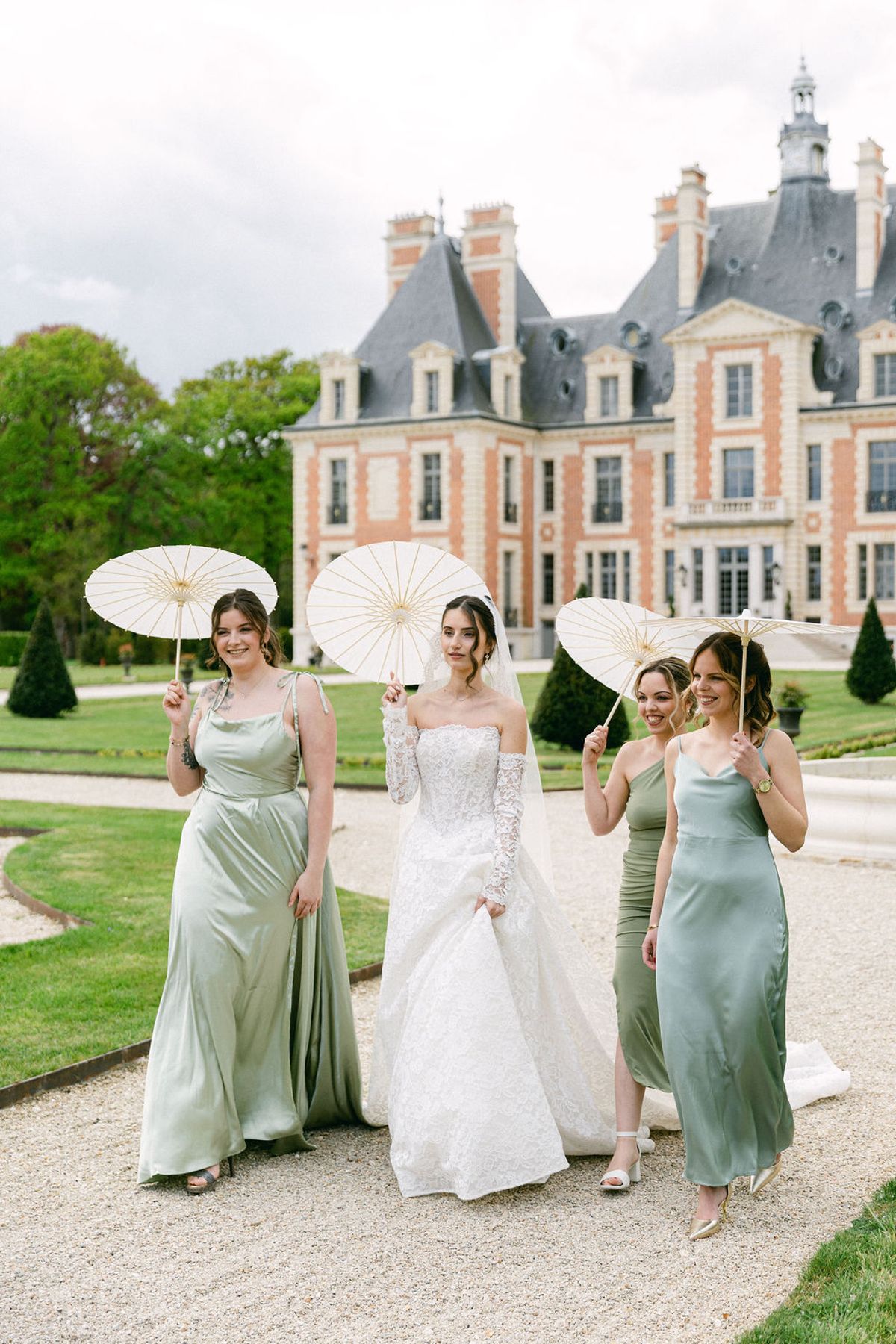 Bride and three bridesmaids in sage green with parasols raised, the chateau behind them