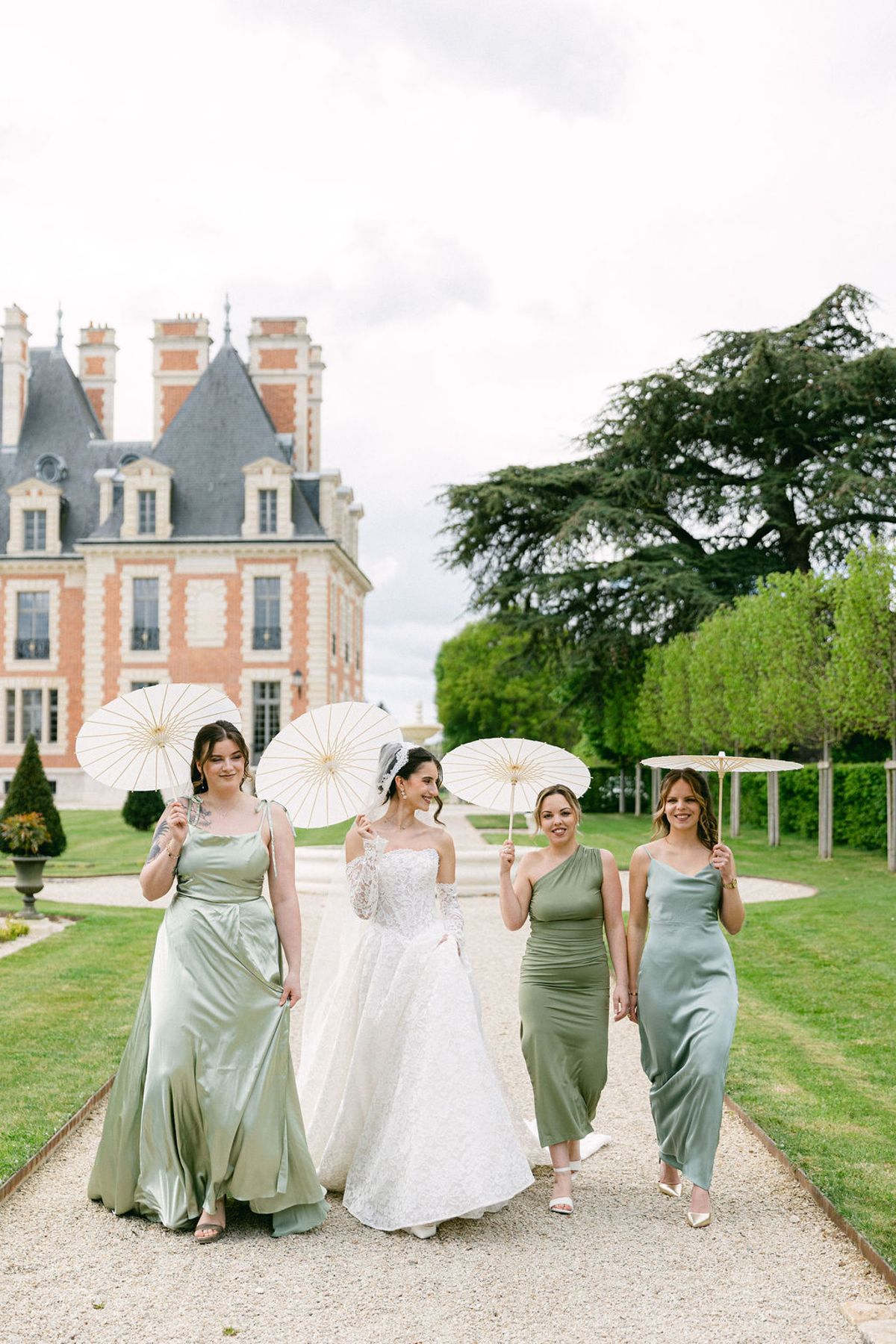 Bride and three bridesmaids in sage green walk down the gravel path carrying parasols