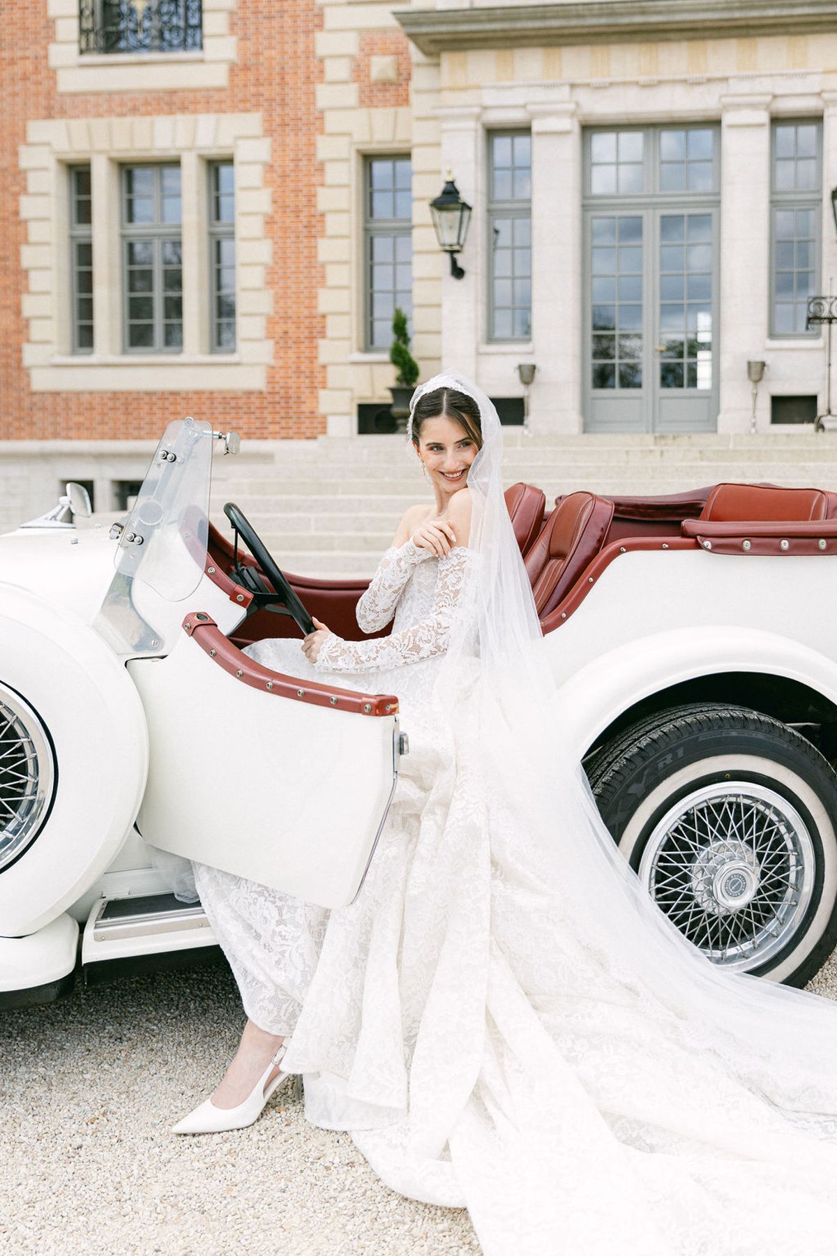 Bride in a lace gown stands beside a white vintage car in front of the chateau