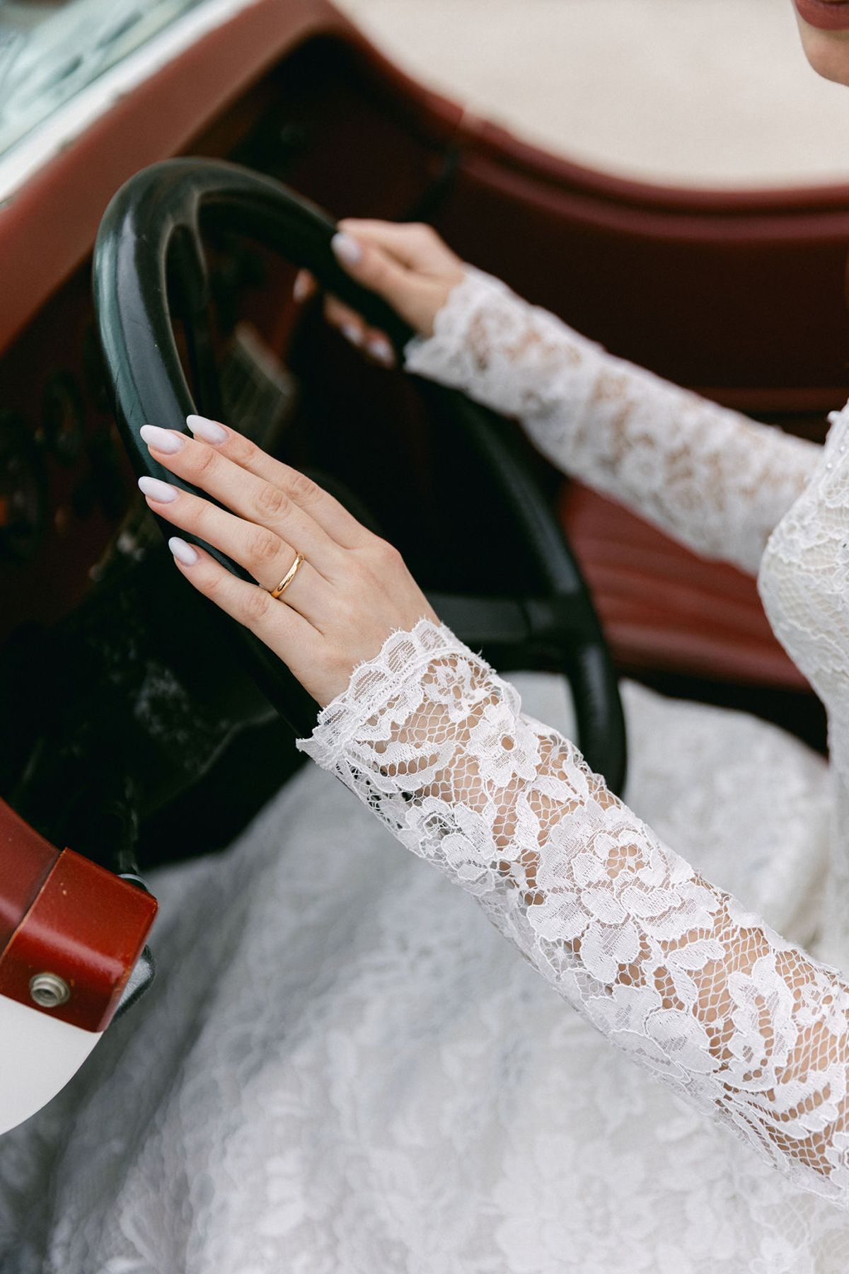 The bride's hands in long lace sleeves resting on the steering wheel of the vintage car