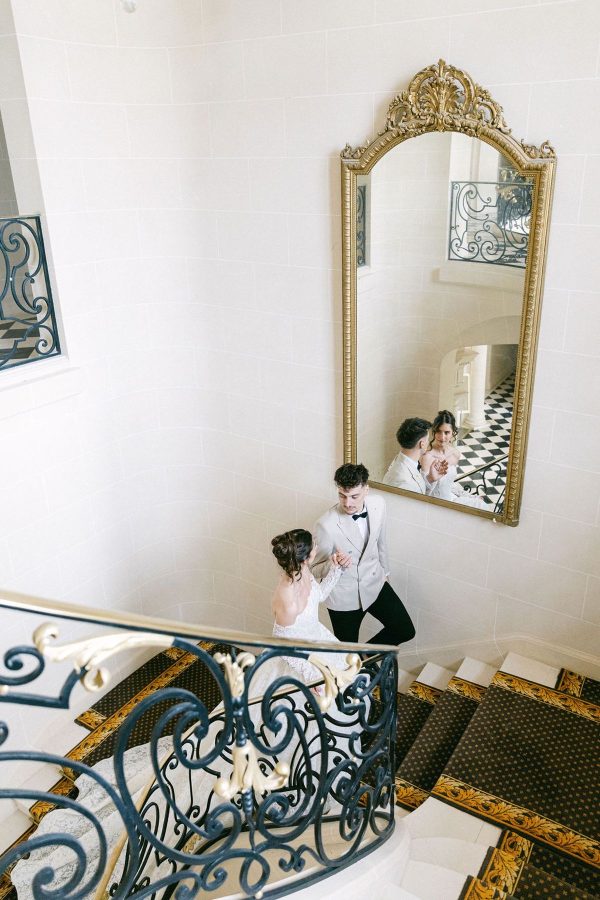 Looking down the chateau stairwell at the couple beside a gilt mirror in the hall below