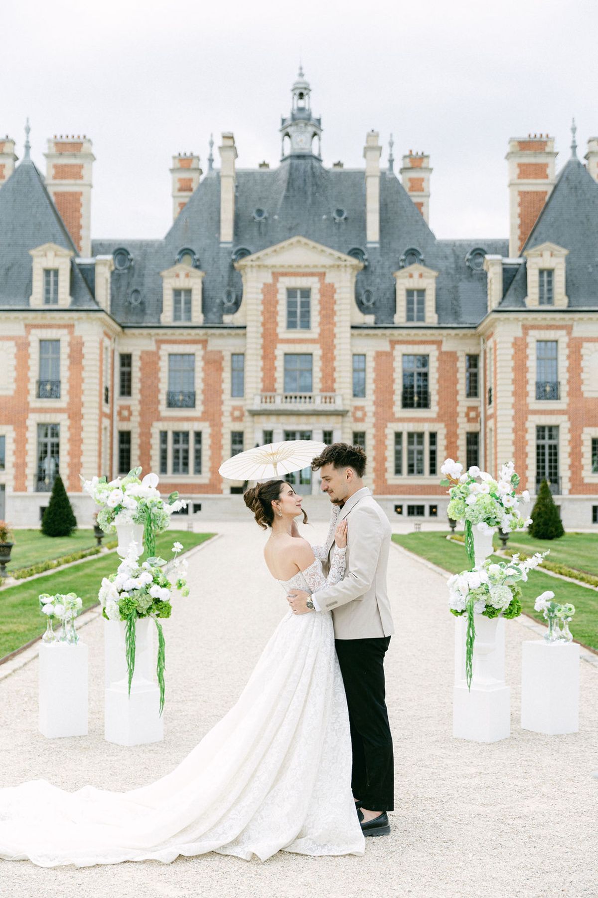 Couple under a parasol on the gravel aisle between the ceremony's floral plinths